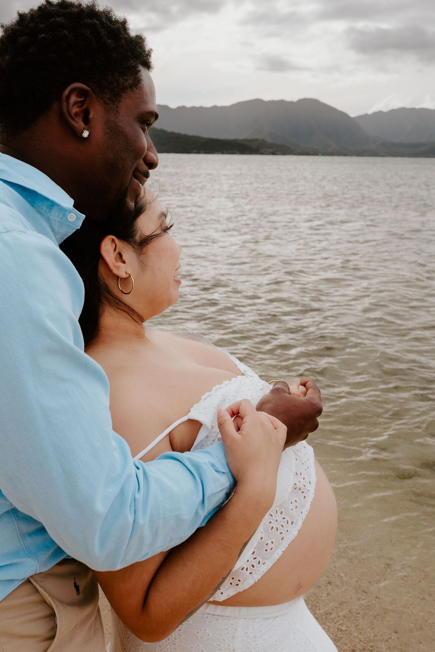 Man embracing pregnant woman from behind while gazing at ocean view.