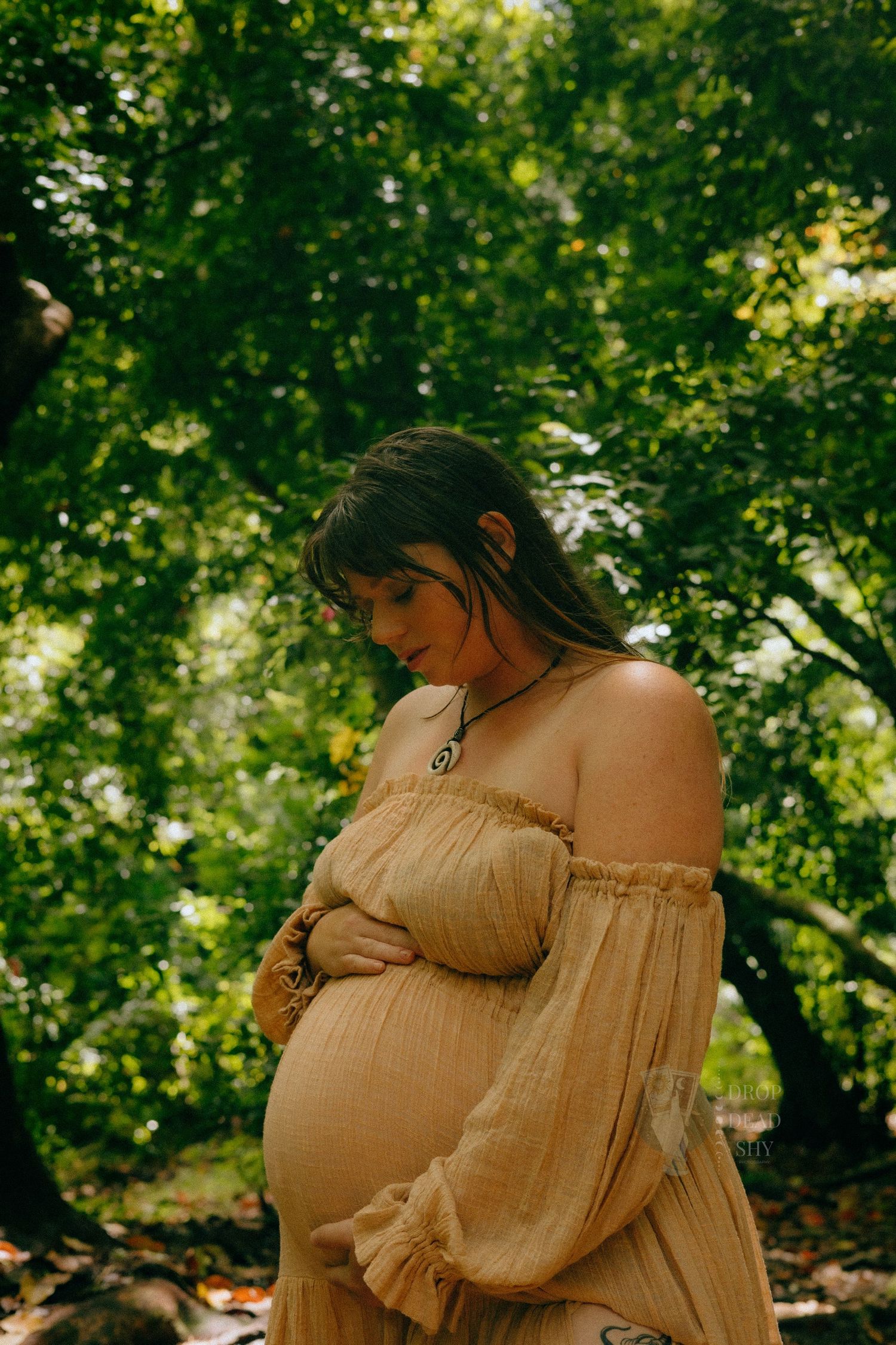 Pregnant woman in golden boho dress tenderly cradling baby bump in forest.
