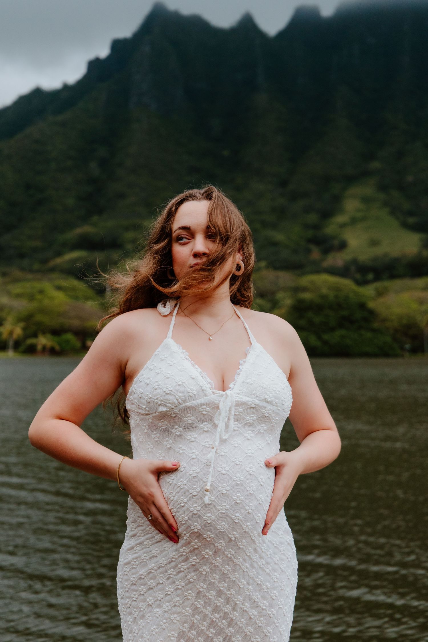 Pregnant woman in white halter dress cradling baby bump near mountain lake in Hawaii.
