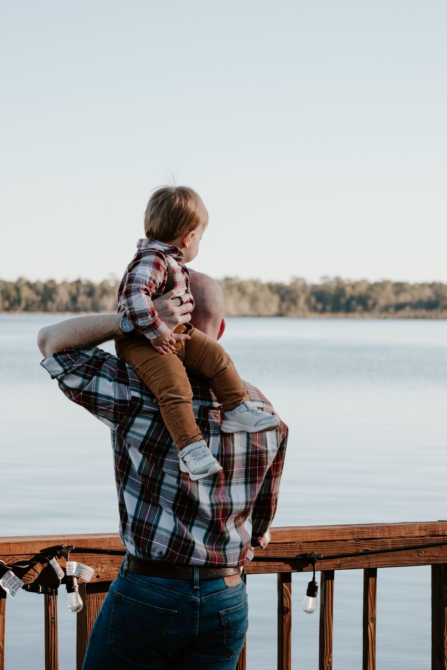 Father lifts toddler onto shoulders while standing on a wooden deck overlooking a calm lake at sunset.