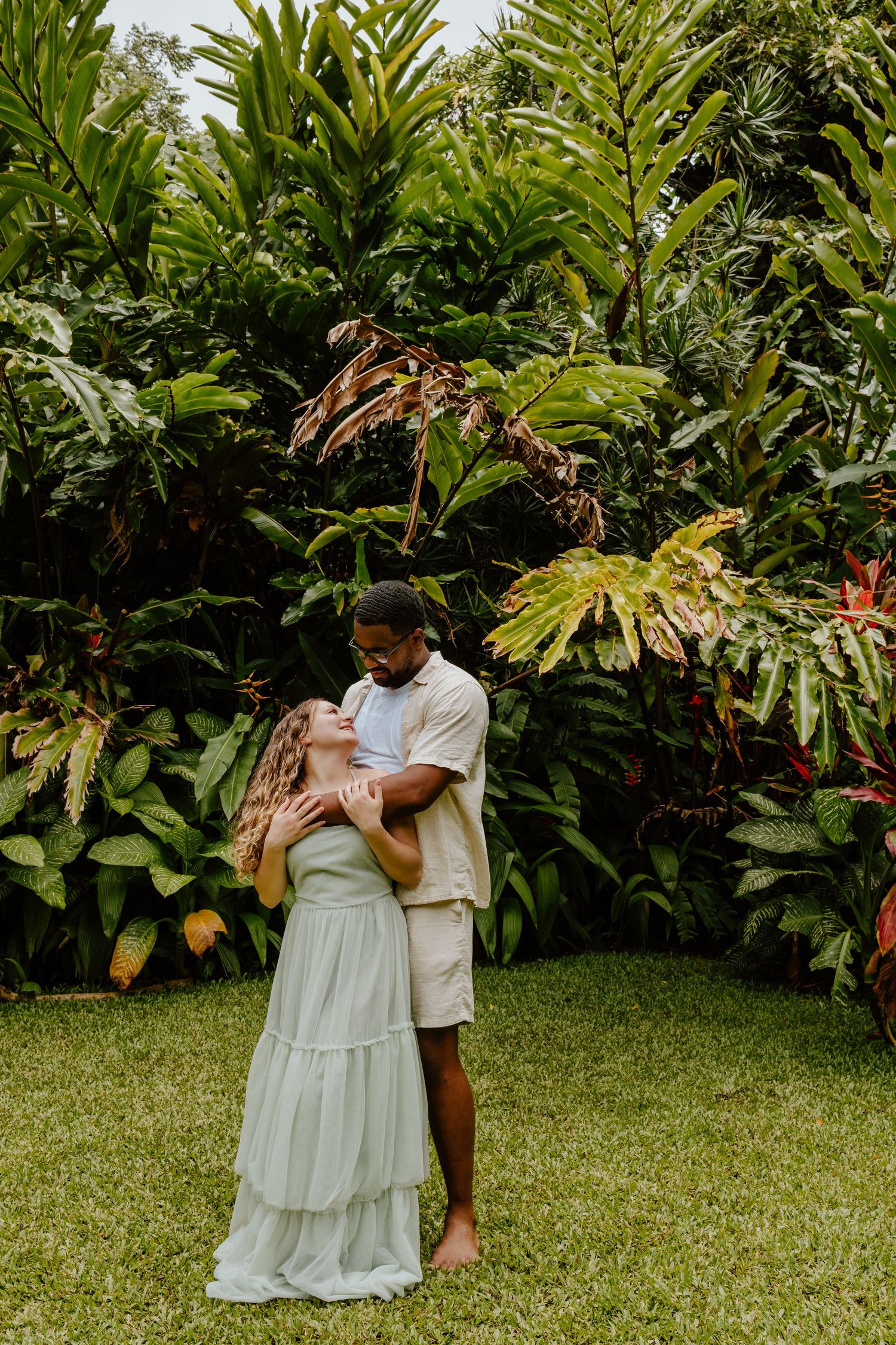 Couple embracing in a lush tropical garden, wearing light neutral outfits, surrounded by vibrant green foliage.