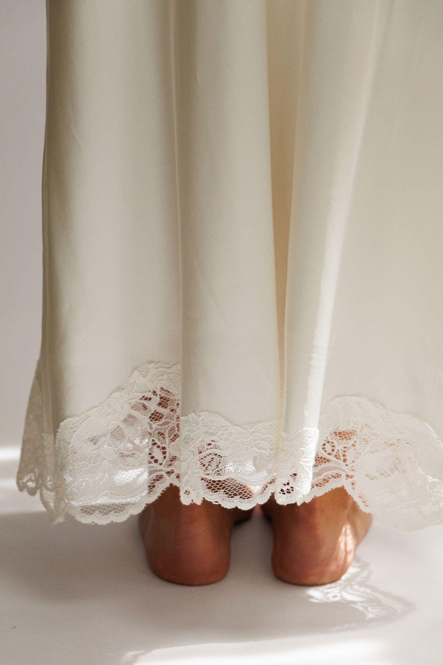 White lace-trimmed slip dress hem with brown heels on white floor.