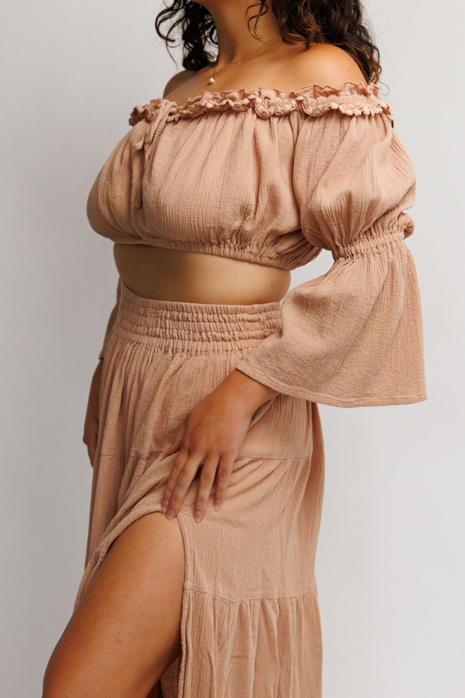 Woman wearing a sandy beige off-shoulder crop top with ruffle trim and matching maxi skirt with side slit.