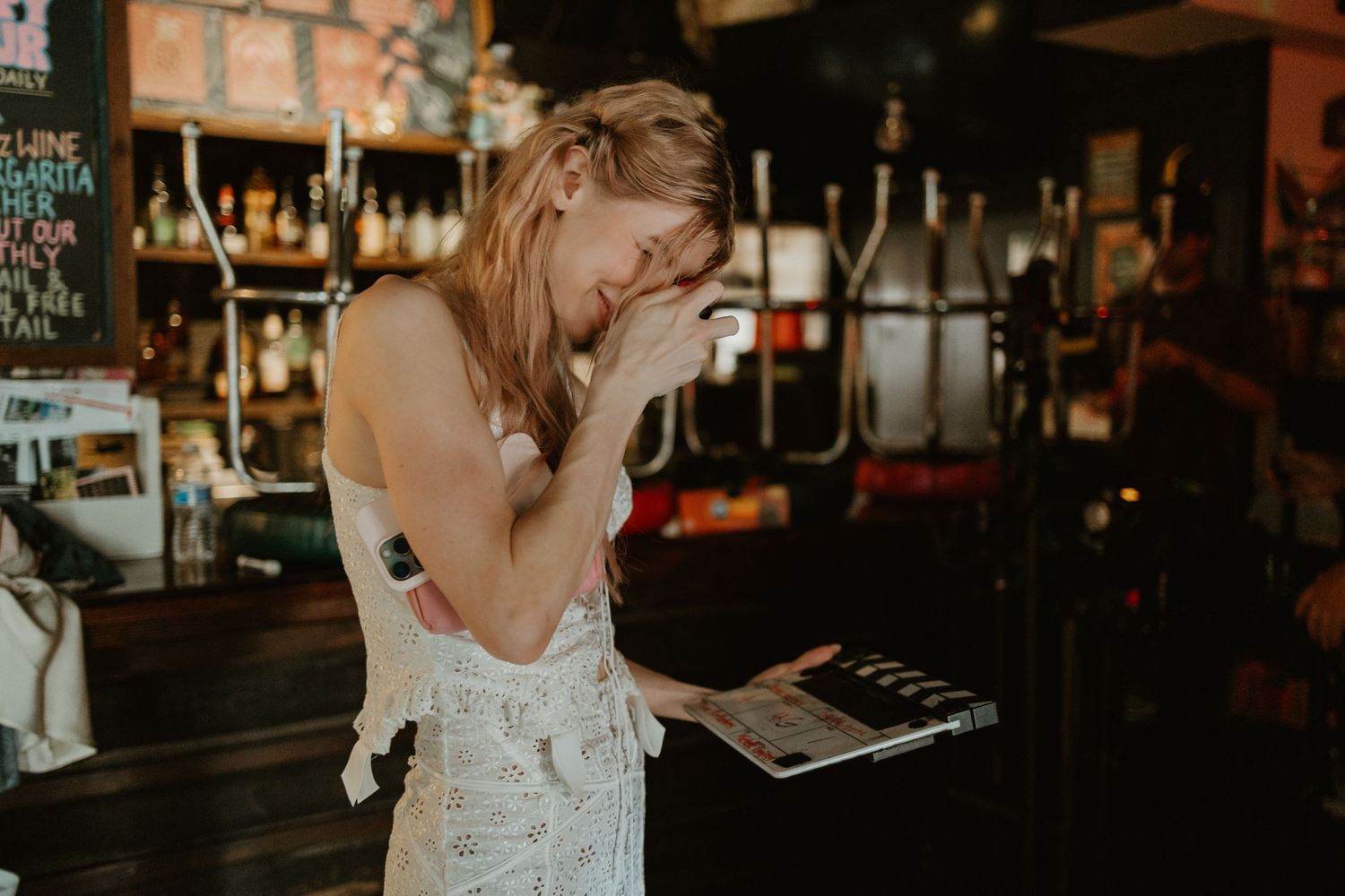 Woman laughing while holding a menu at a bar counter with colorful bottles in the background.