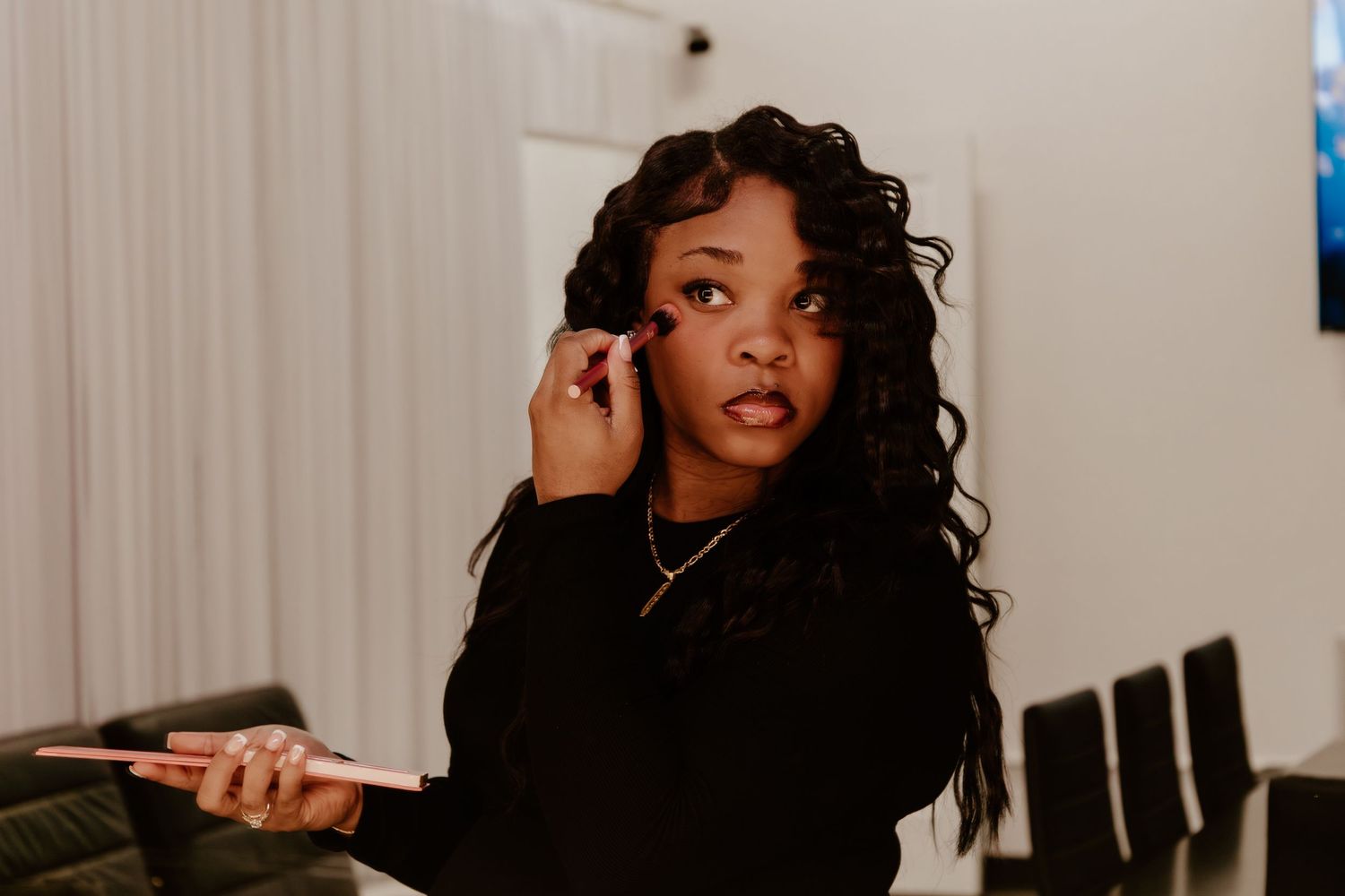 Young Black woman in black outfit applying makeup with a brush, holding a tablet in a bright room.