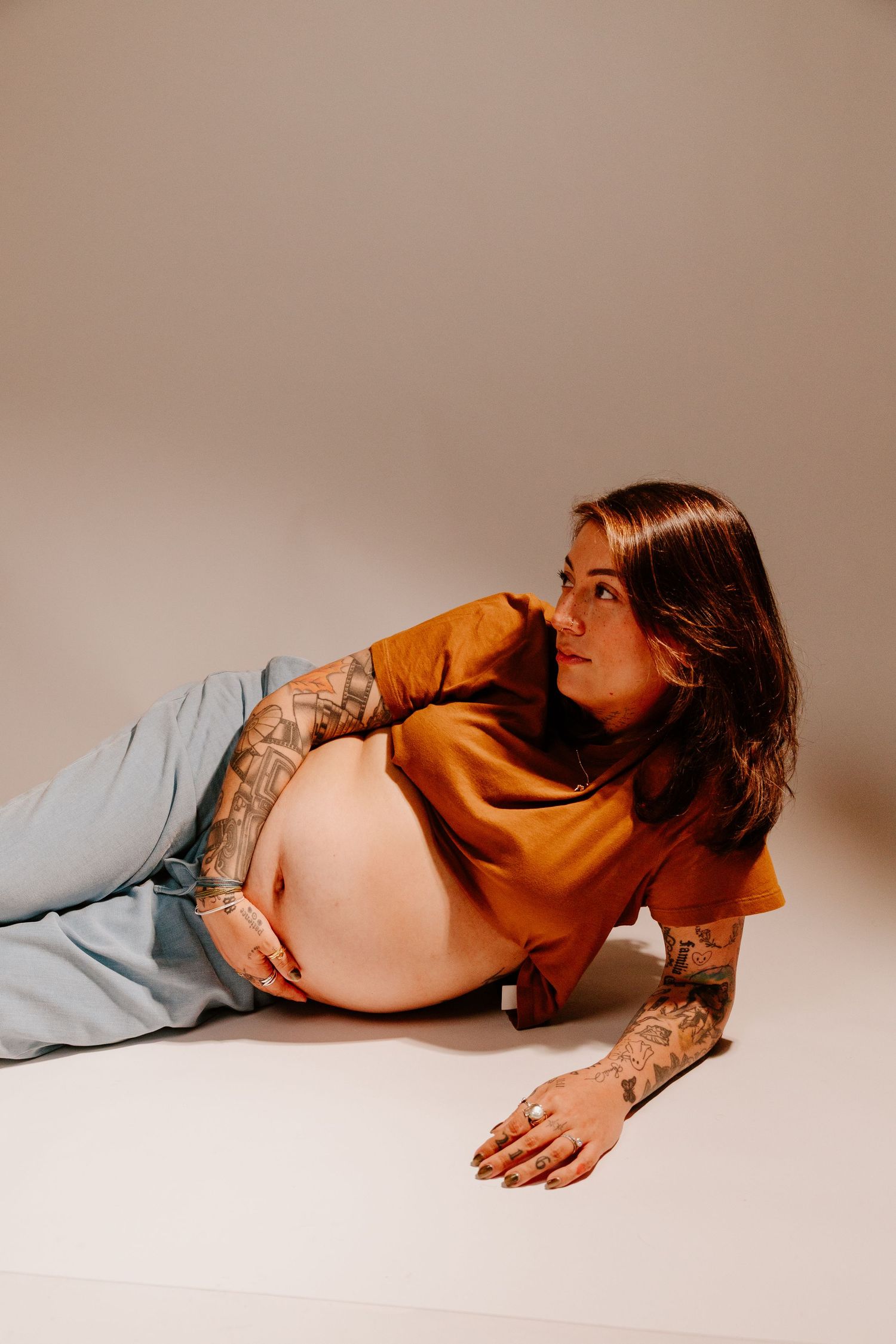 Tattooed pregnant woman in orange shirt lying on floor, gazing to the side.