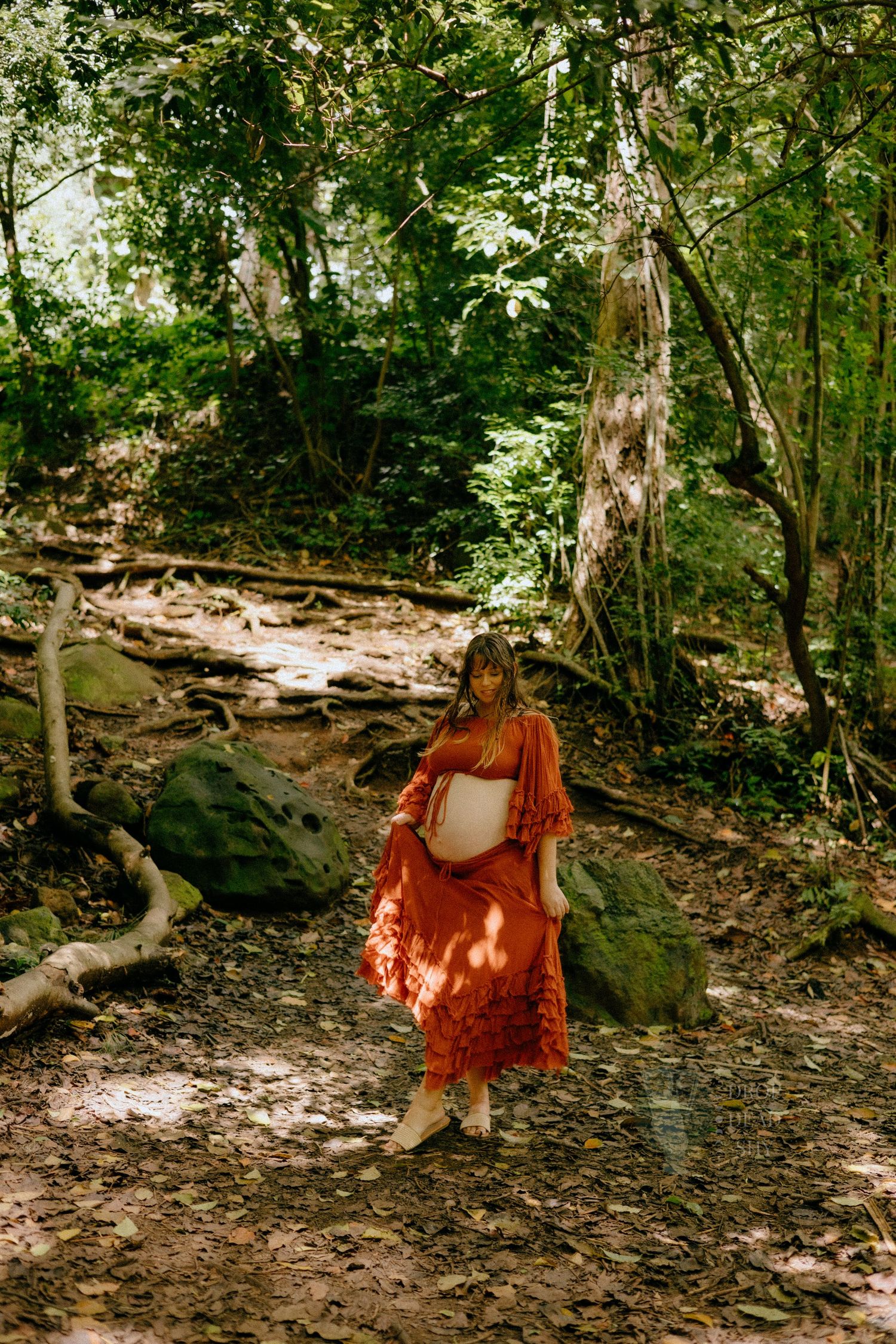 Pregnant woman in rust orange maxi dress cradling belly among mossy forest rocks.