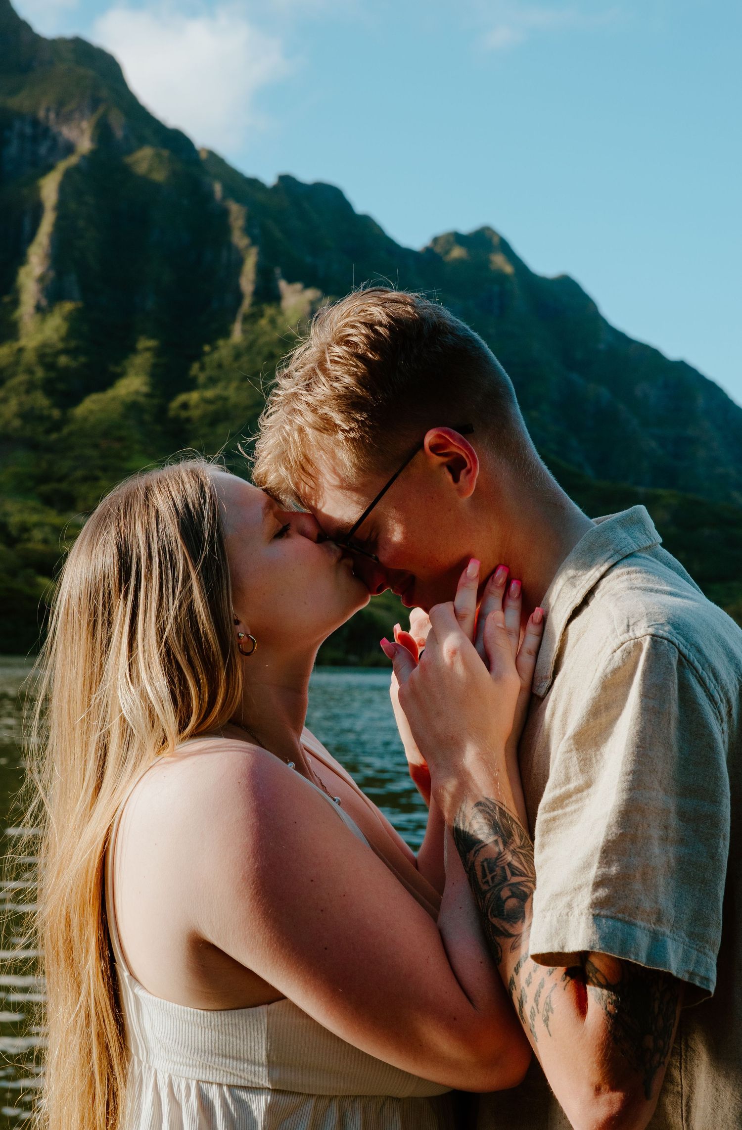 A couple shares a passionate kiss near a mountain lake with lush green peaks in the background.