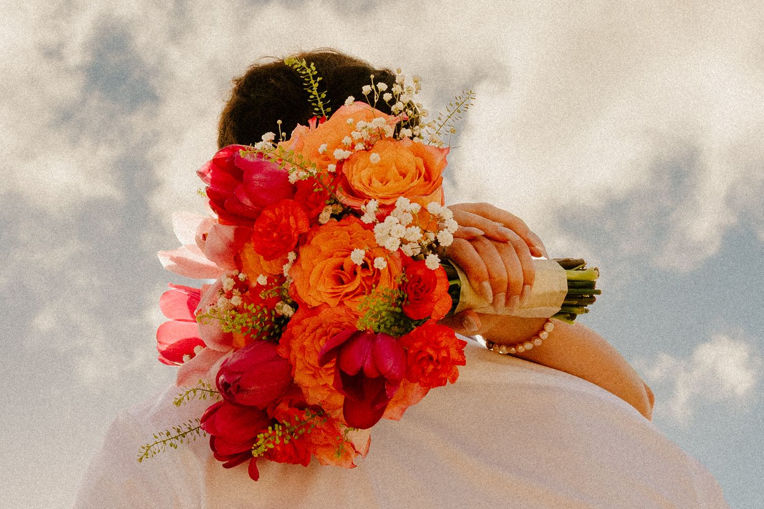 Bride holding a vibrant wedding bouquet of orange roses and red flowers against a cloudy sky.