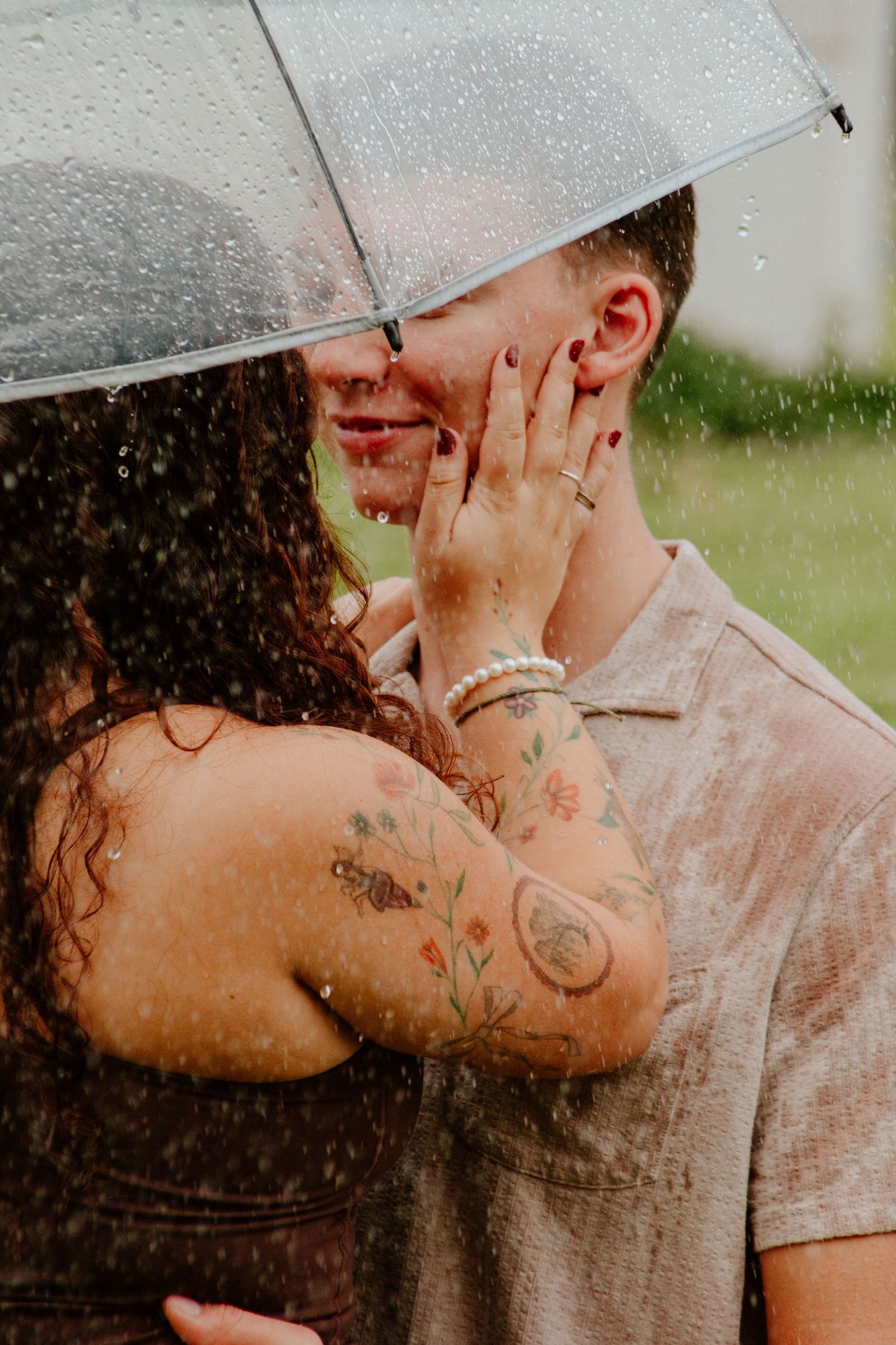 Couple embracing and kissing in the rain under a clear umbrella, woman with colorful floral tattoos.