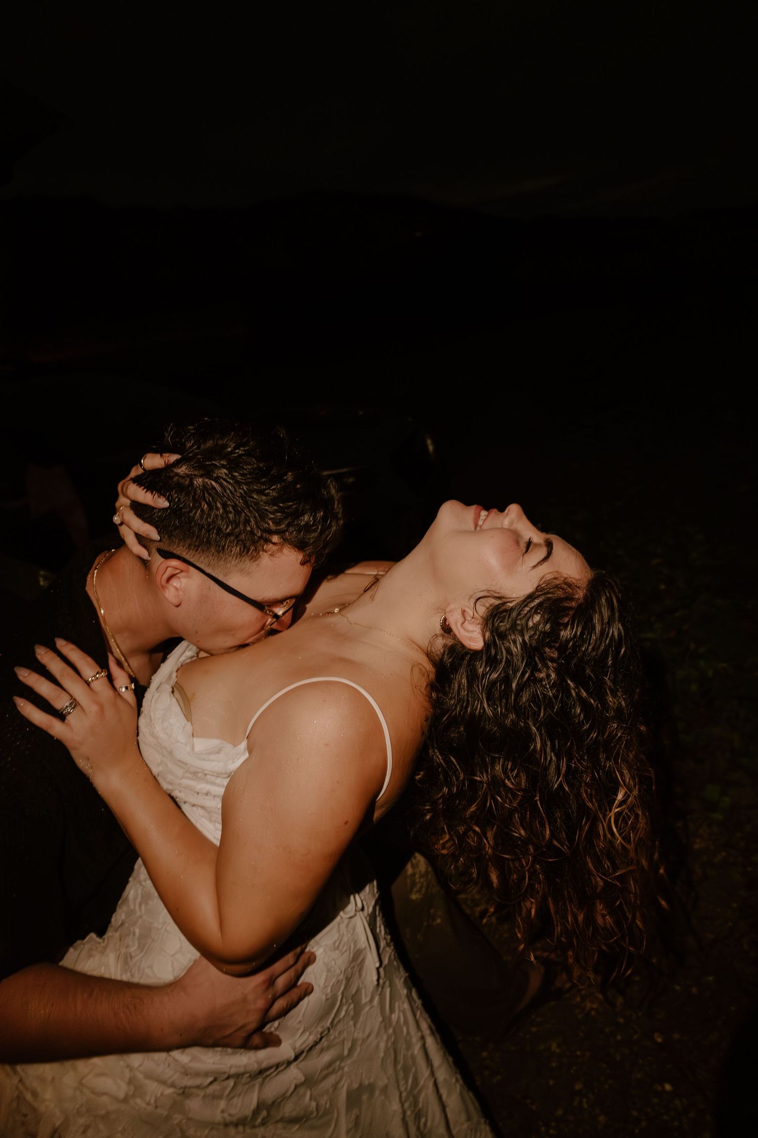 A couple shares a passionate embrace at night, with the bride laughing joyfully in a white dress.