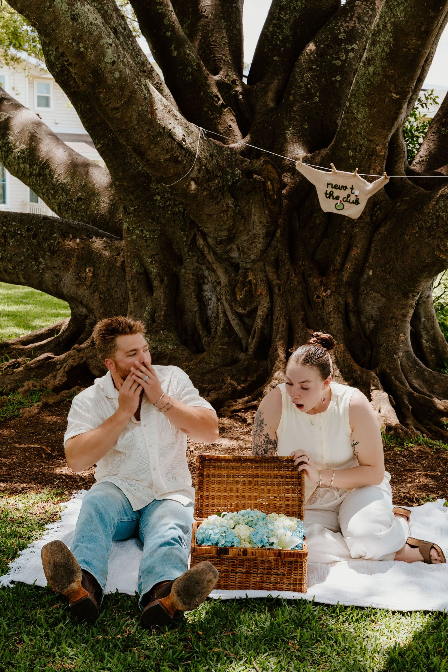 Couple reacting with surprise to blue flowers in wicker basket during gender reveal under tree.