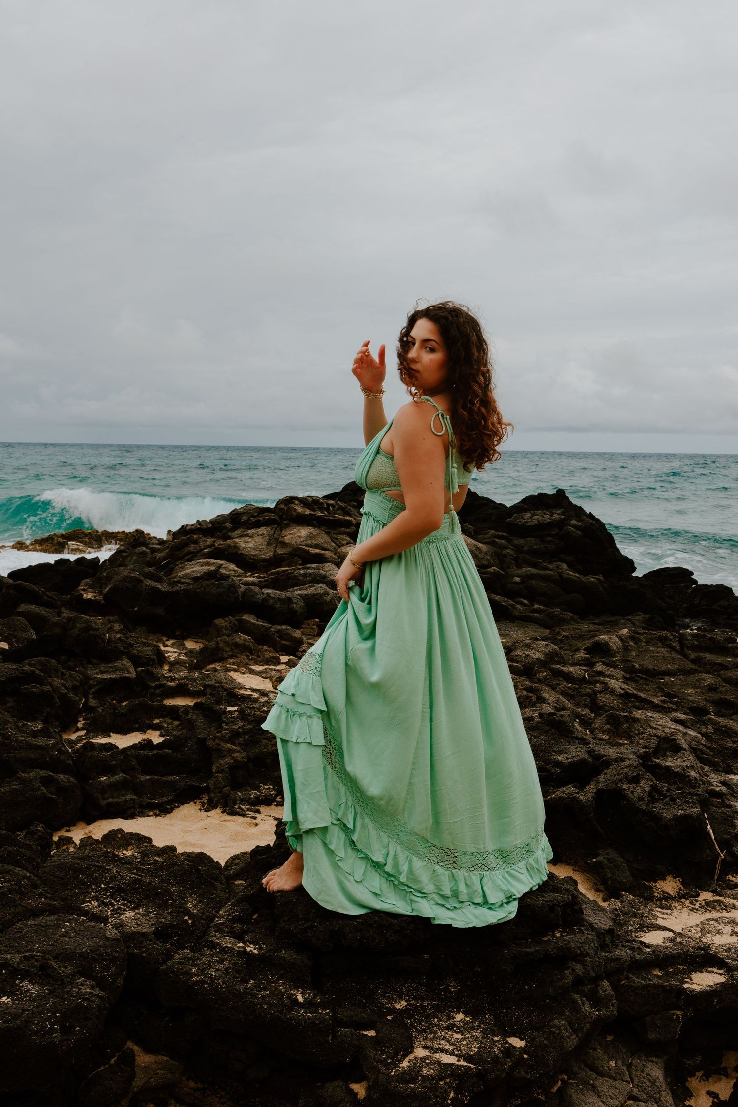 Woman in mint green maxi dress standing on volcanic rocks by turquoise ocean under cloudy sky.