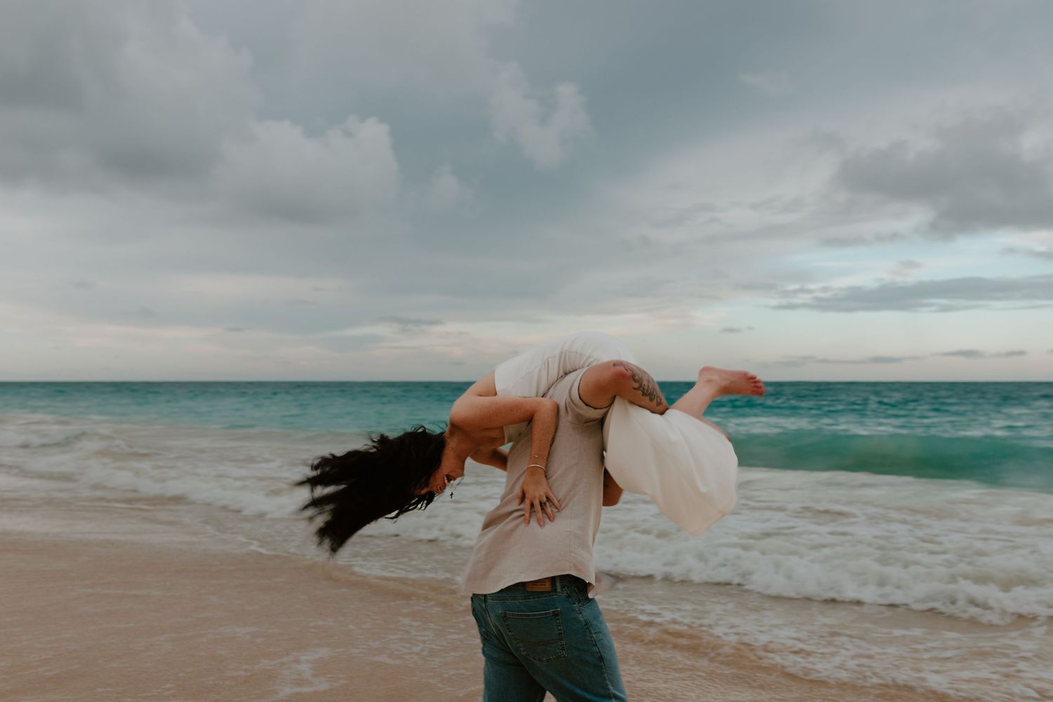 A man lifts a woman in a white dress on a tropical beach with turquoise waves under a cloudy sky.
