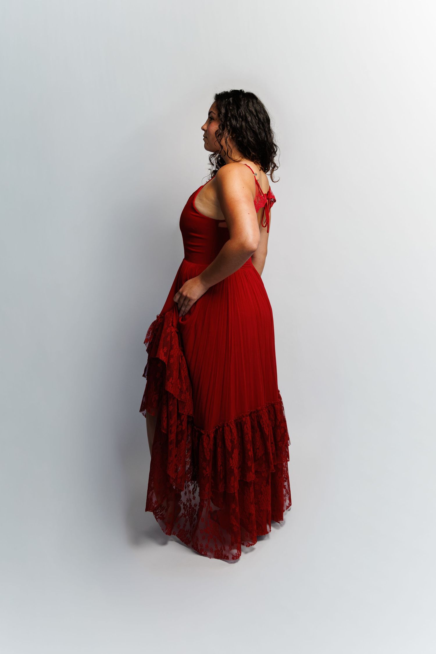 Woman in elegant red lace maxi dress with ruffled skirt, side profile view, studio photography.