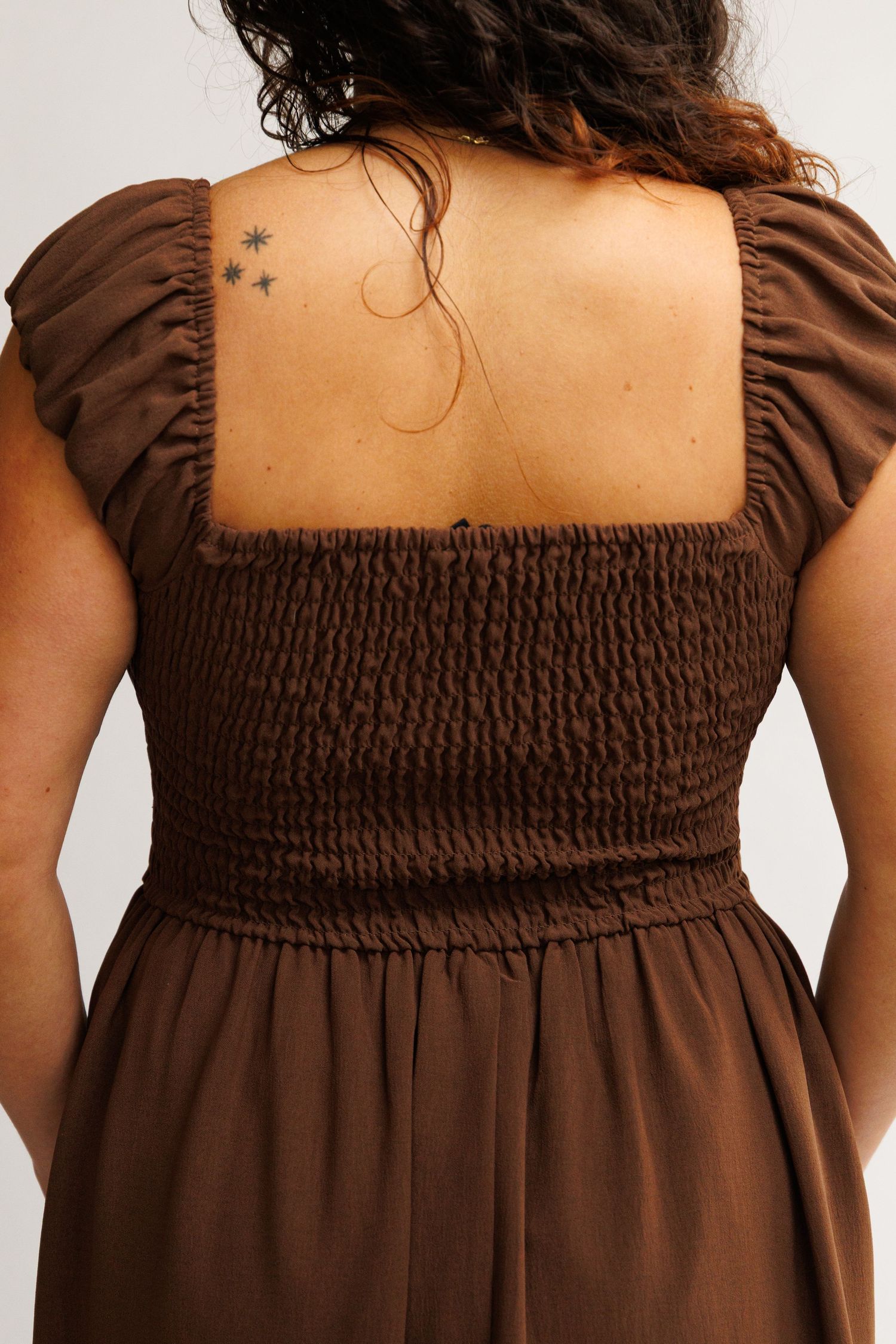 Back view of a brown smocked bodice dress with puff sleeves and square neckline, worn by a model with star tattoos.