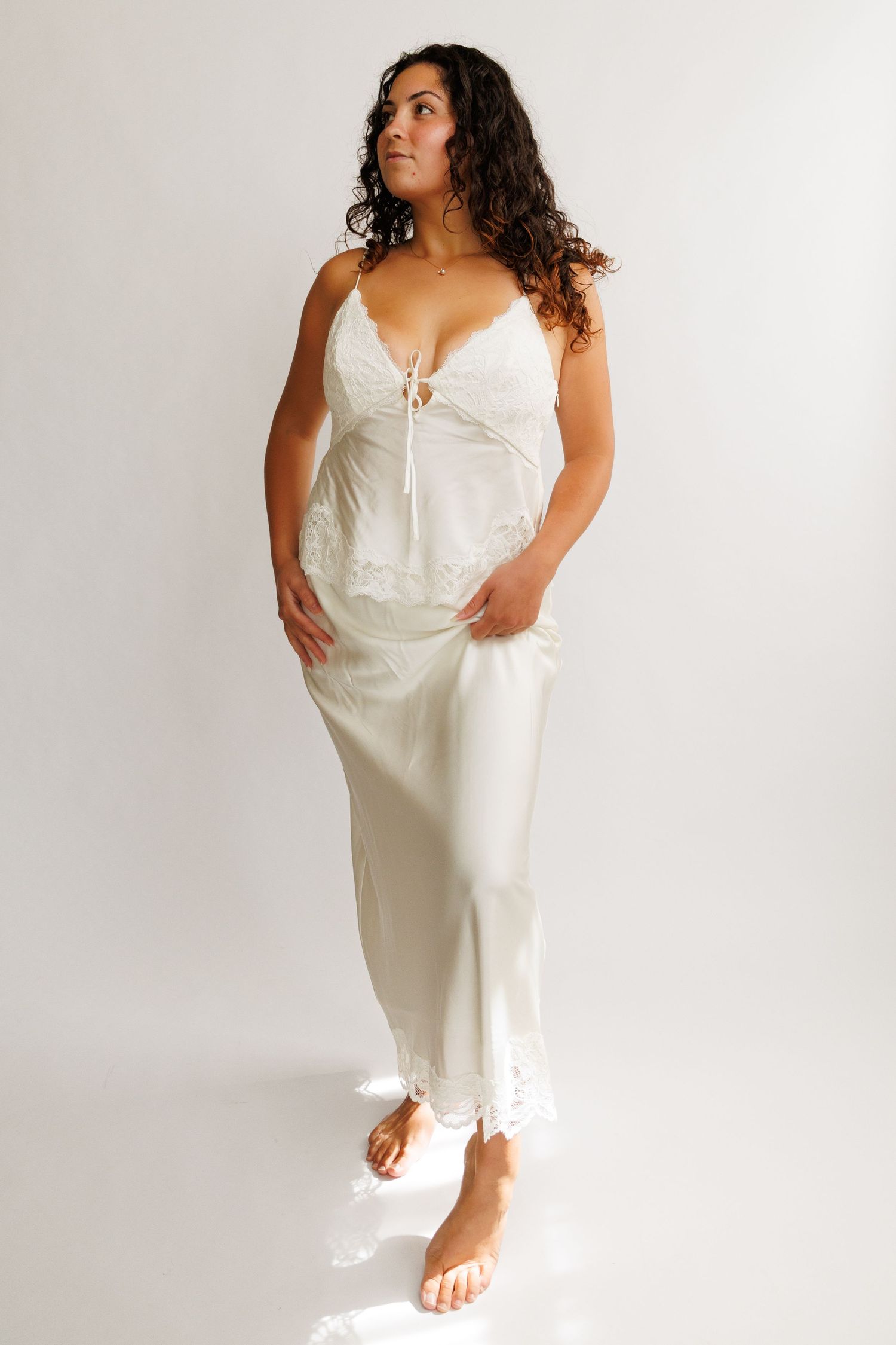Woman wearing white lace camisole top and satin midi skirt with lace hem, standing barefoot on white background.