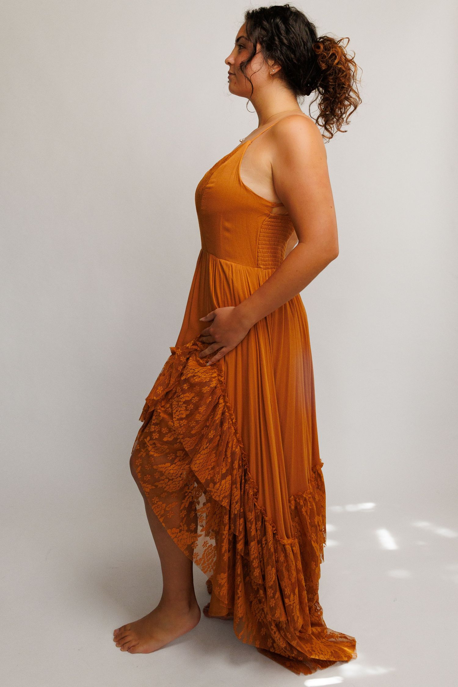 Woman wearing a burnt orange halter maxi dress with lace hem detail, posing sideways in a studio setting.