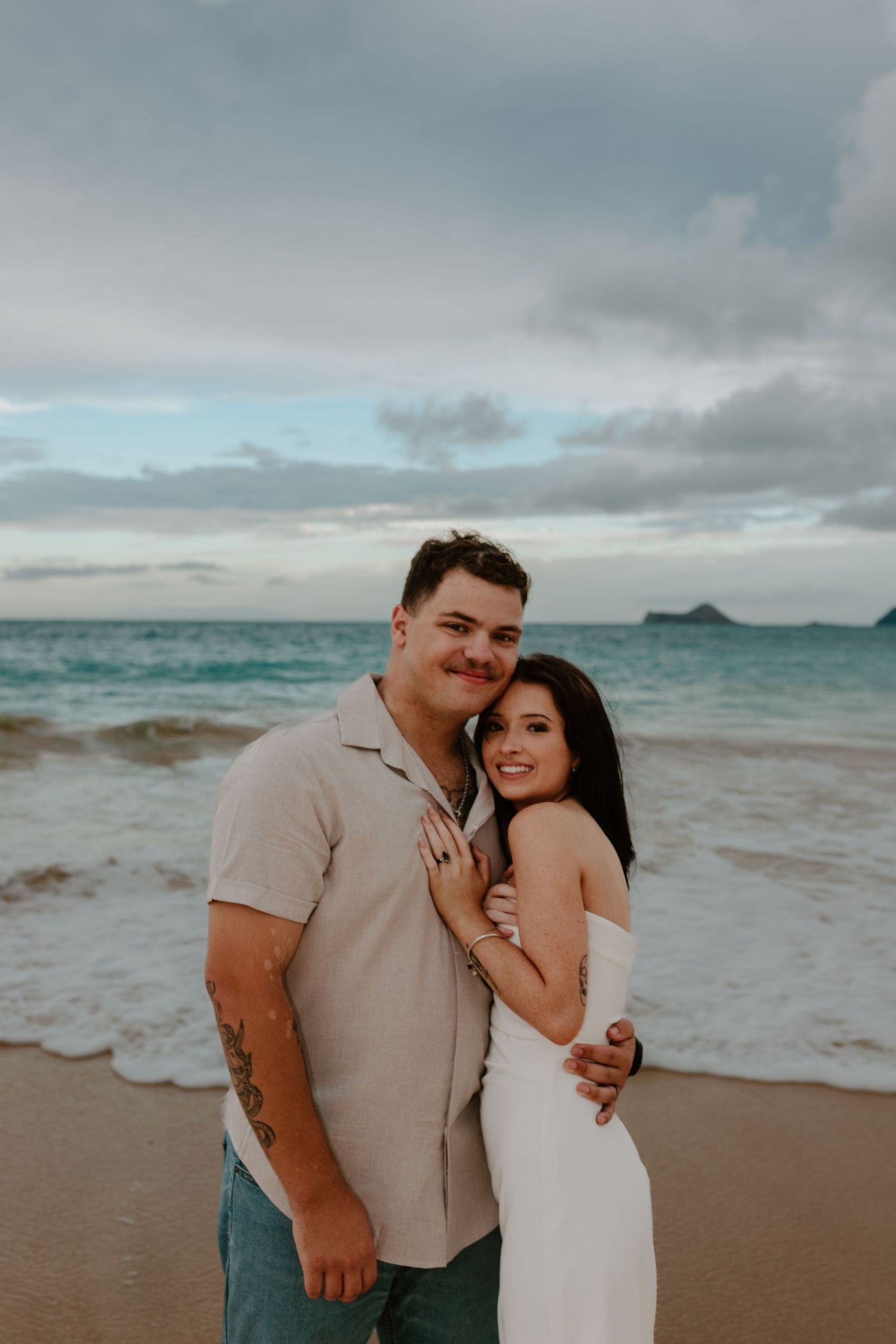 Smiling couple posing on a Hawaiian beach at dusk, woman in white dress embracing tattooed man in beige shirt.