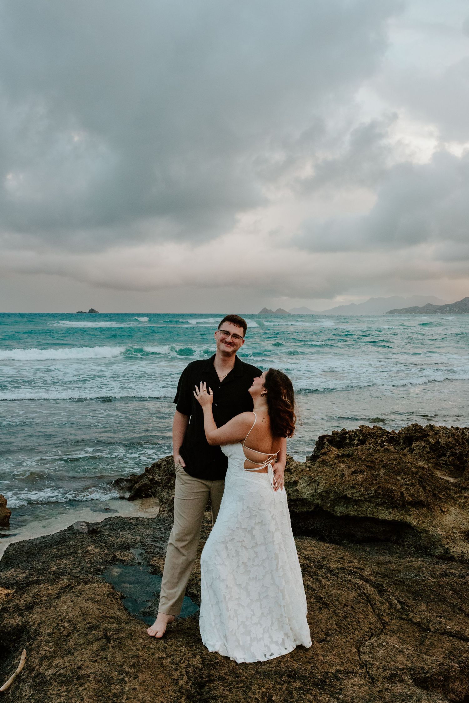 A smiling couple in wedding attire embraces on rocky shore with turquoise ocean waves in the background.