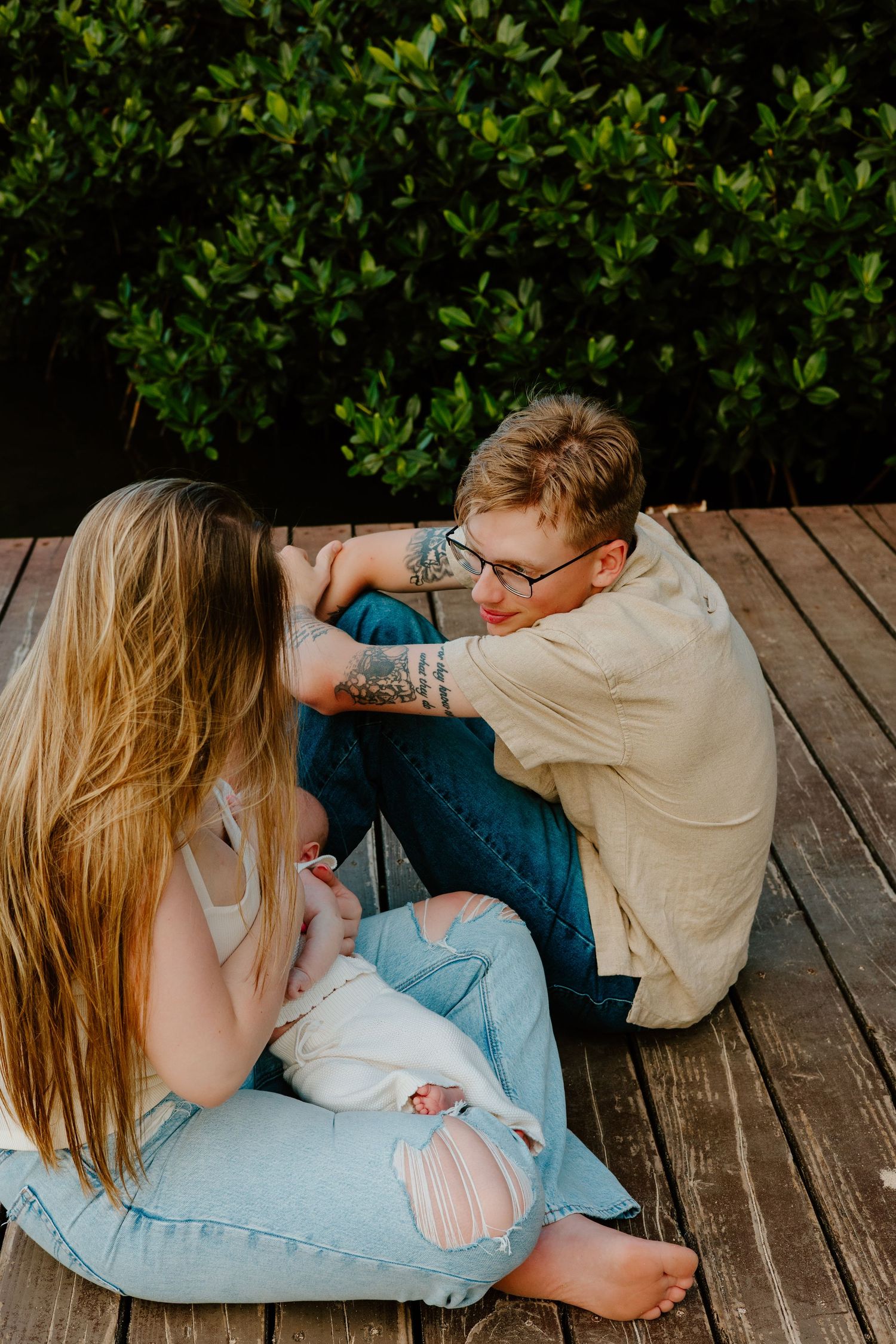 Young couple with baby sitting on wooden deck surrounded by lush green foliage.