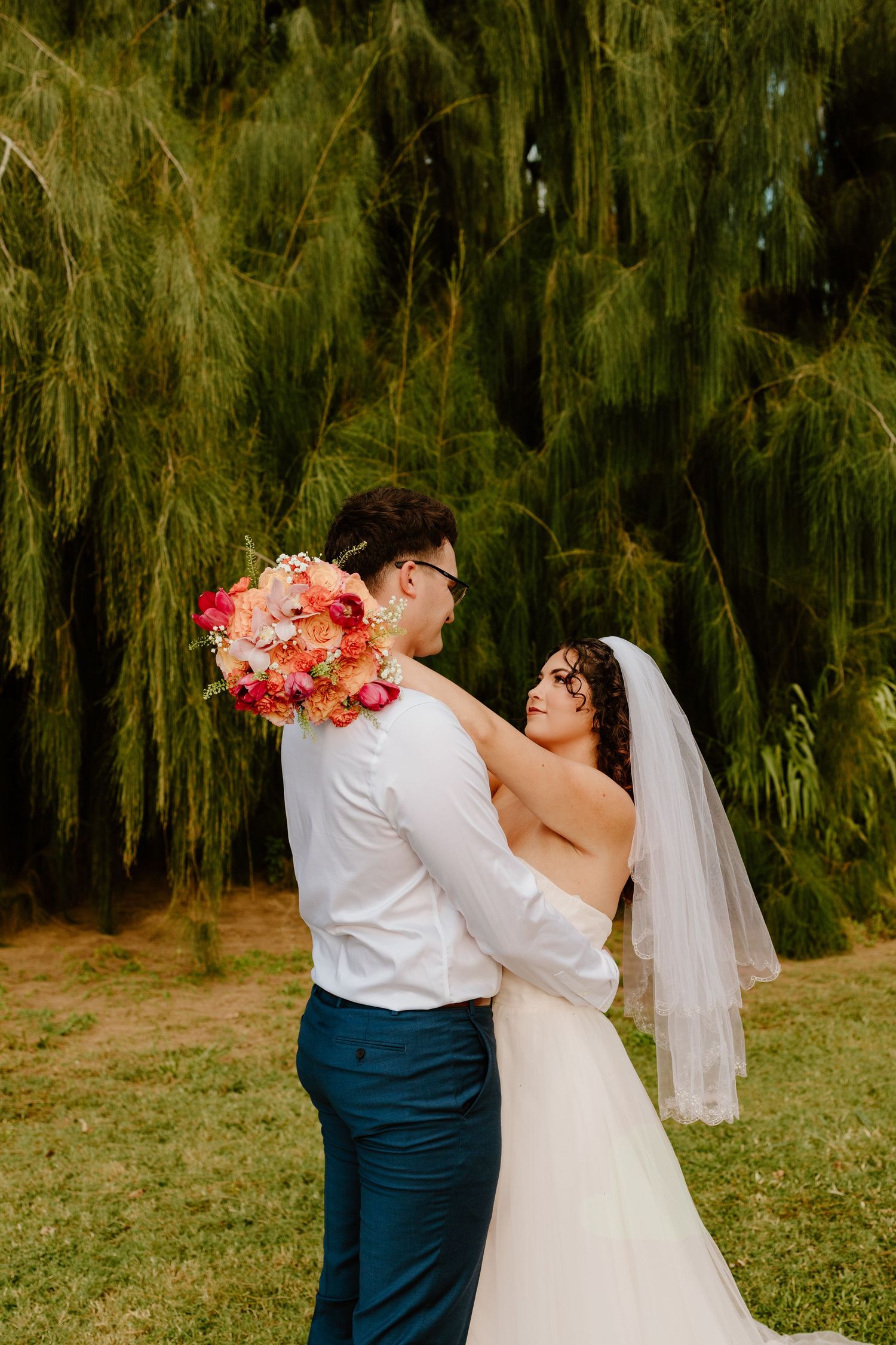 Bride and groom embracing outdoors under a weeping willow tree, bride holding a vibrant pink and red floral bouquet.