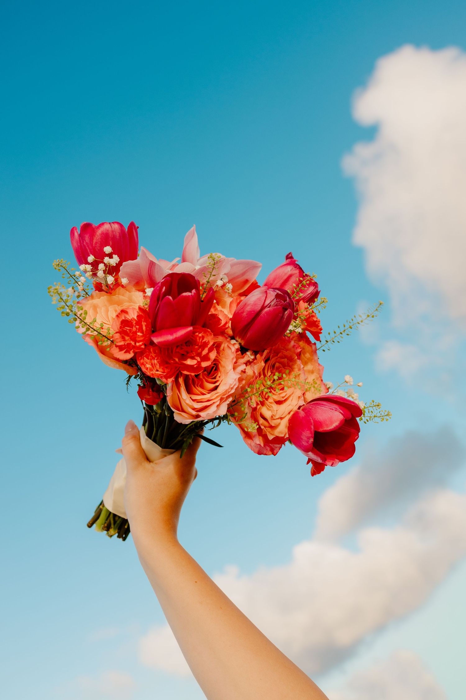 Hand holding a vibrant bouquet of red tulips and pink roses raised against a bright blue sky with white clouds.