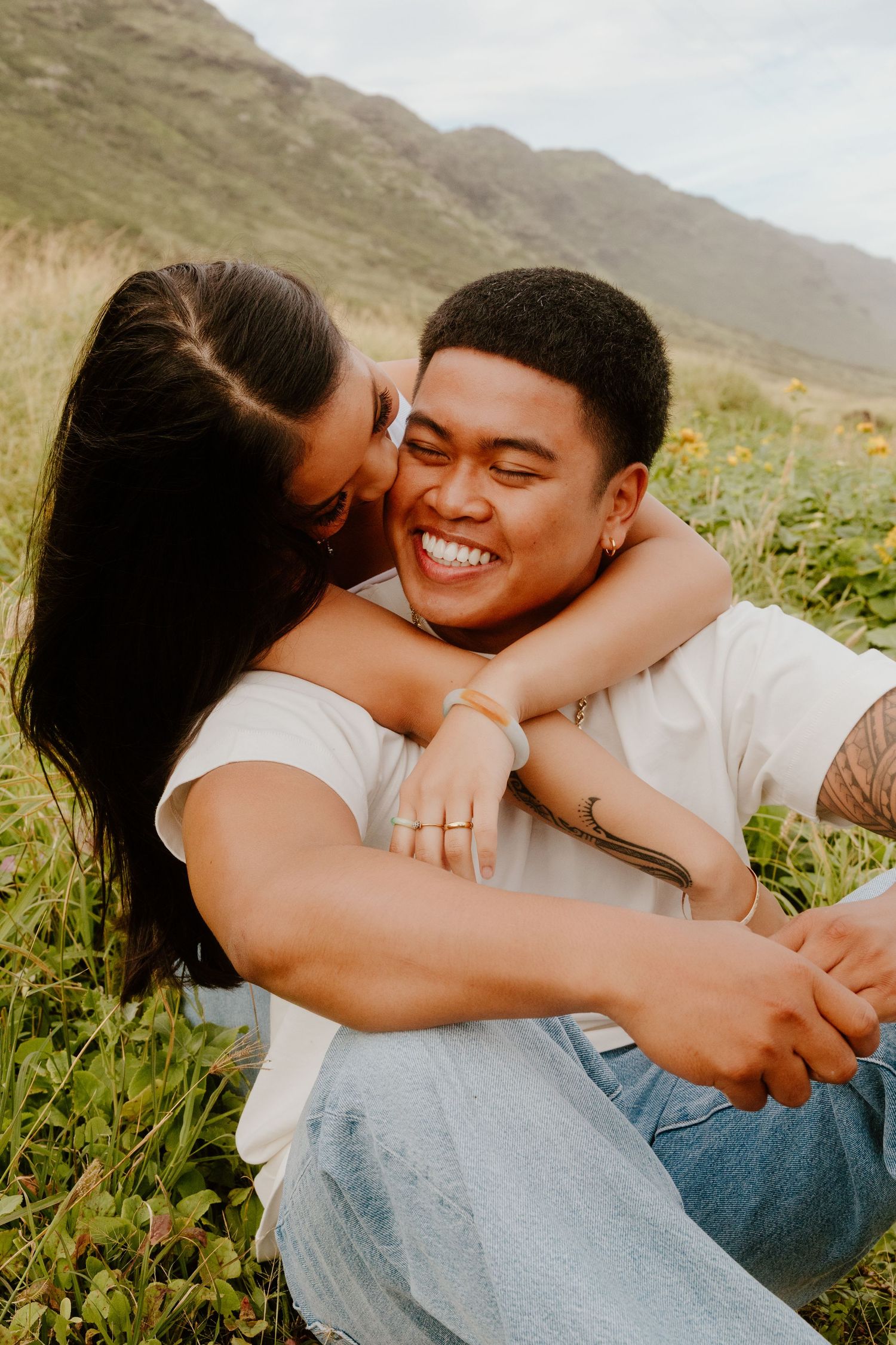 A joyful couple sits in a grassy field, the woman kissing the mans cheek as he laughs happily.