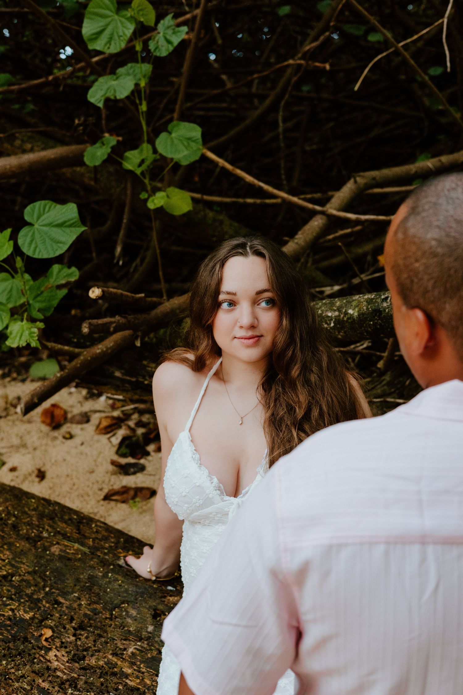 Young woman in a white lace dress looking up at her partner on a sandy tropical beach setting.