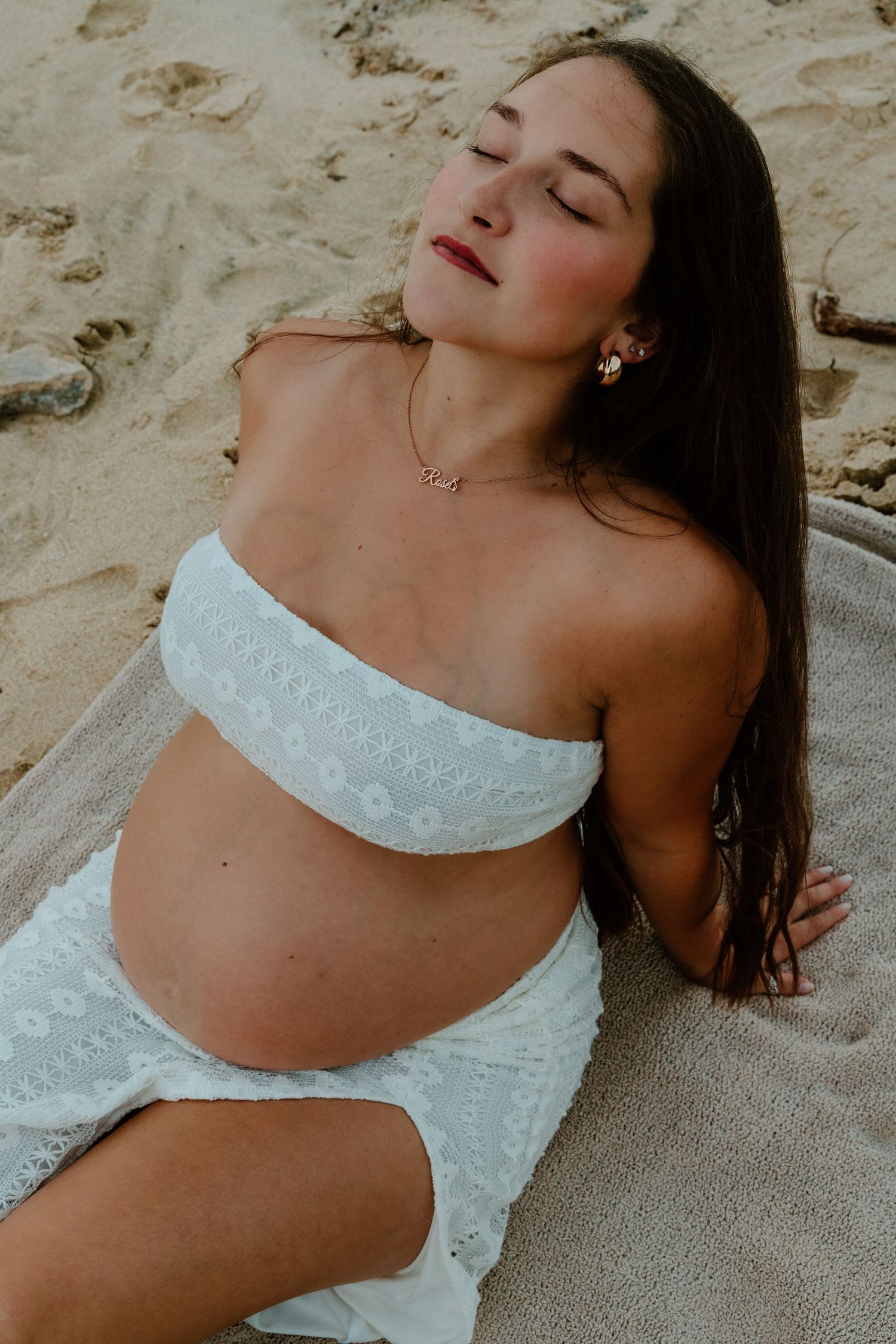 Serene pregnant woman in white bandeau top relaxing on sandy beach.