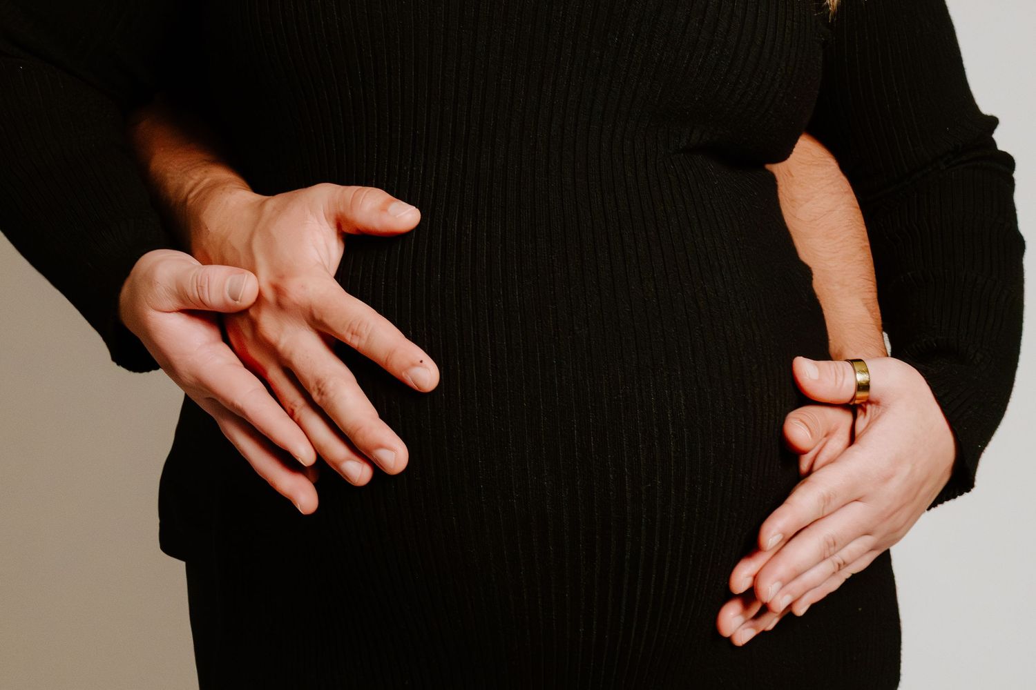 Close-up of pregnant woman in black dress cradling baby bump with both hands, wearing gold ring.