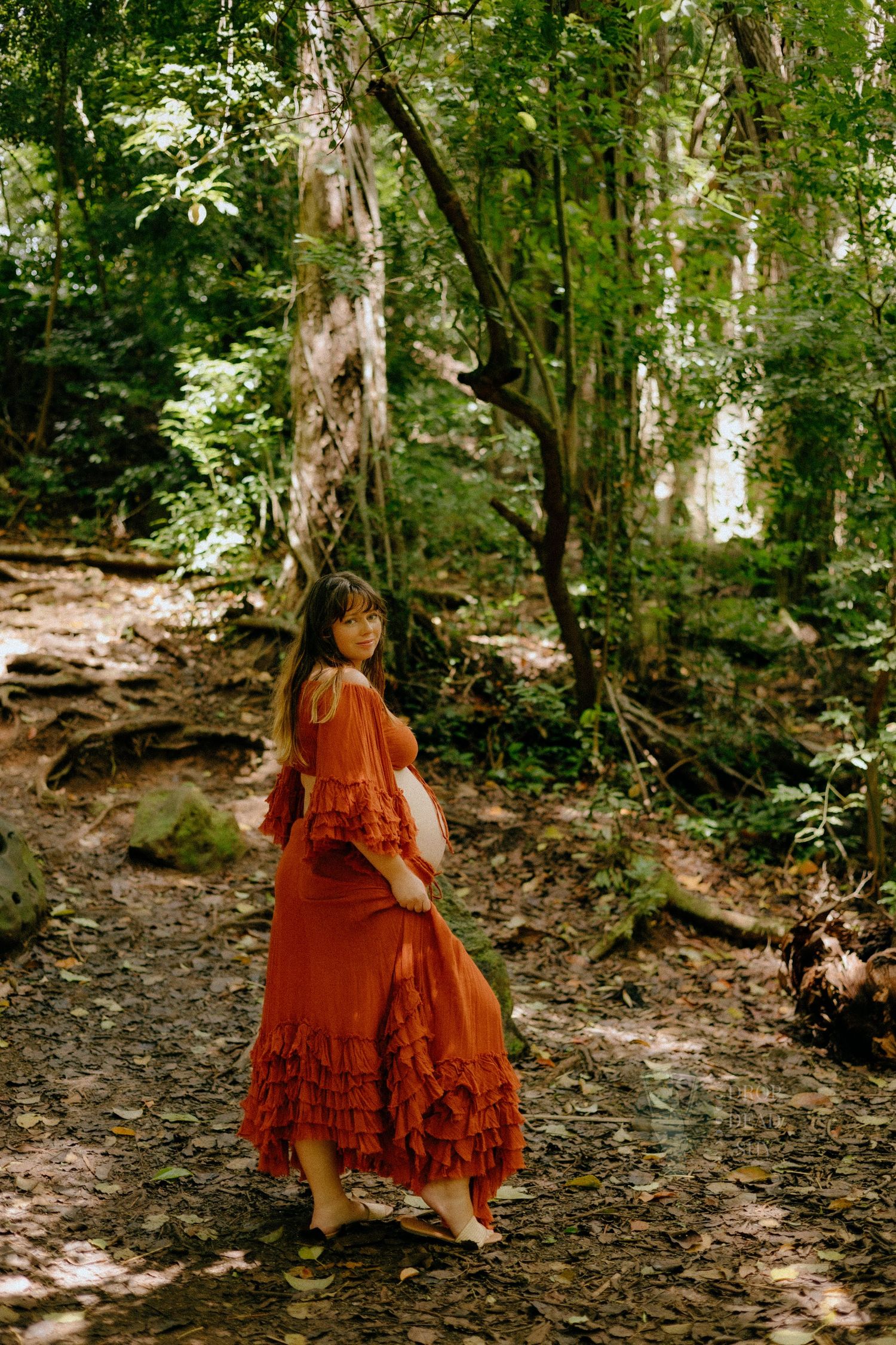 Pregnant woman in a flowing rust-red ruffled dress standing on a forest path surrounded by lush green trees.