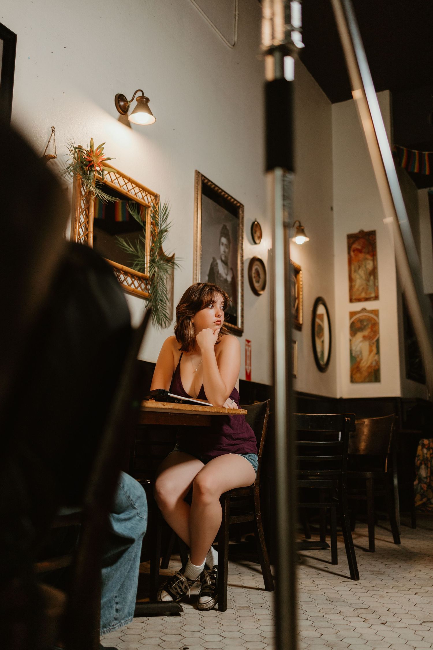 Woman sitting alone at a wooden table in a vintage cafe, looking thoughtful with framed artwork on walls.