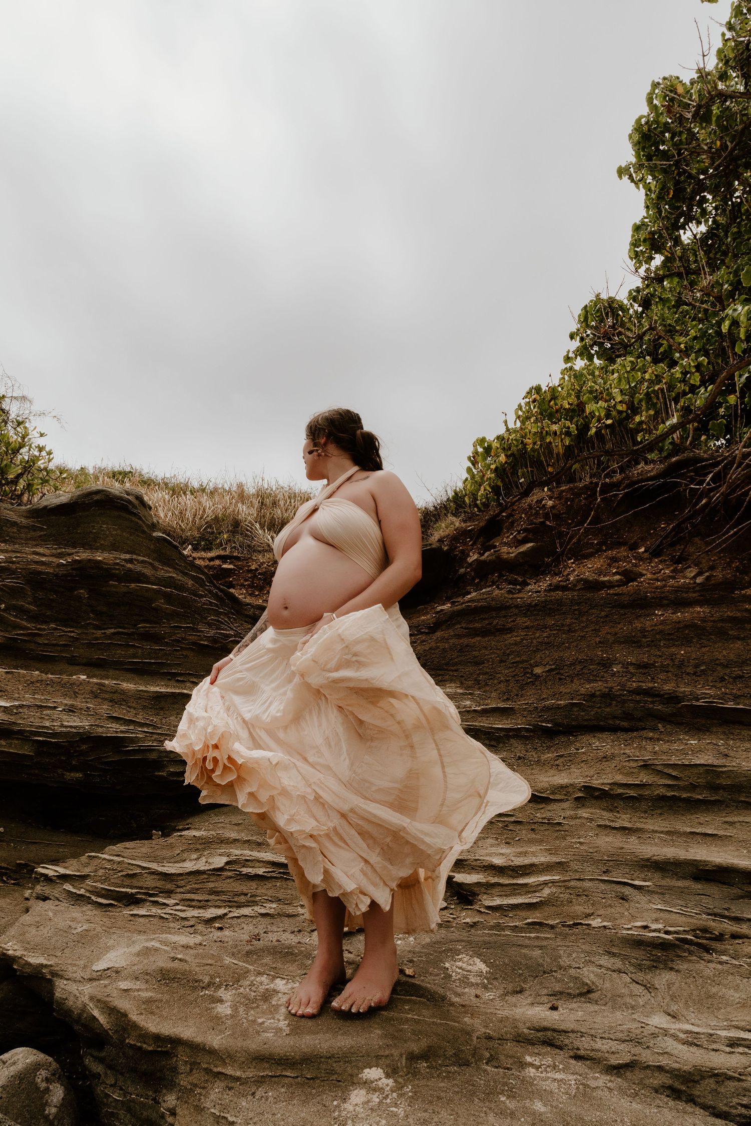 Pregnant woman in flowing peach gown standing barefoot on rocky coastal cliffs with lush greenery backdrop.