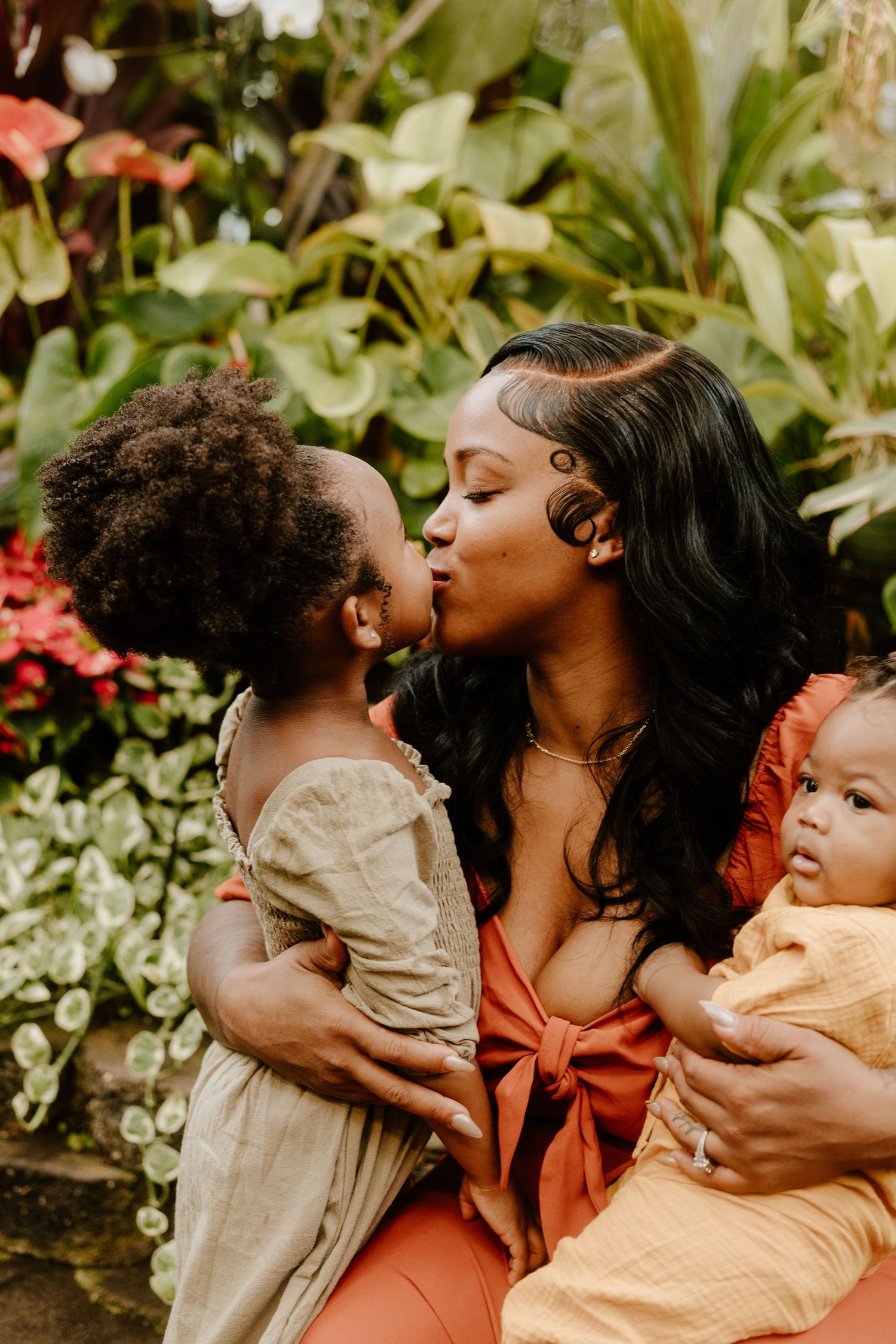 A mother kisses her toddler while holding her baby surrounded by tropical plants.
