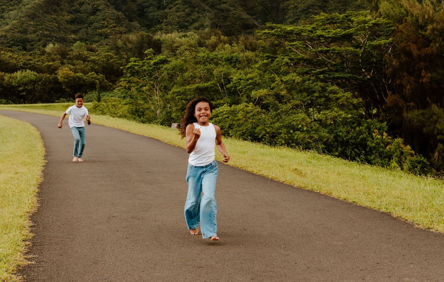 Girl in white top and jeans running on a scenic road surrounded by lush green trees.
