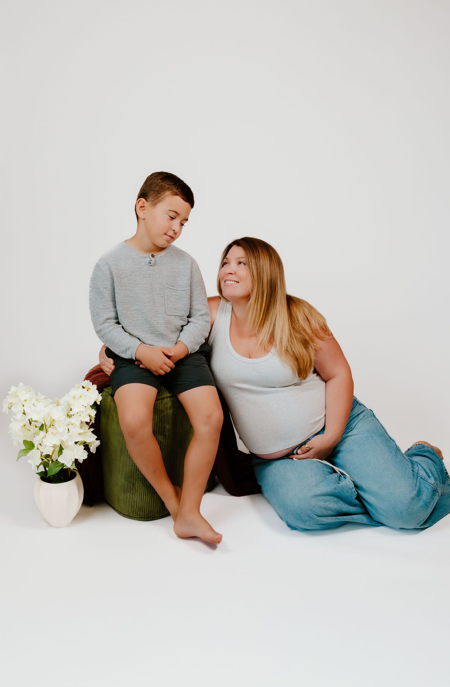 Pregnant woman in jeans and tank top sits with young boy on green ottoman beside white flowers on white background.