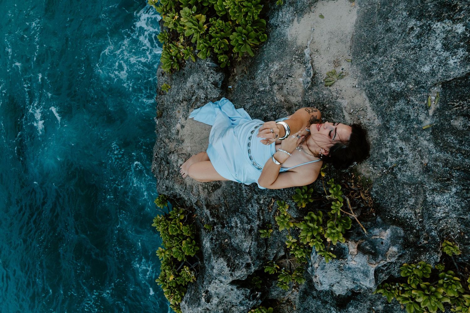 Aerial view of a woman in a white dress lying on rocky coastal cliffs above turquoise ocean waters.