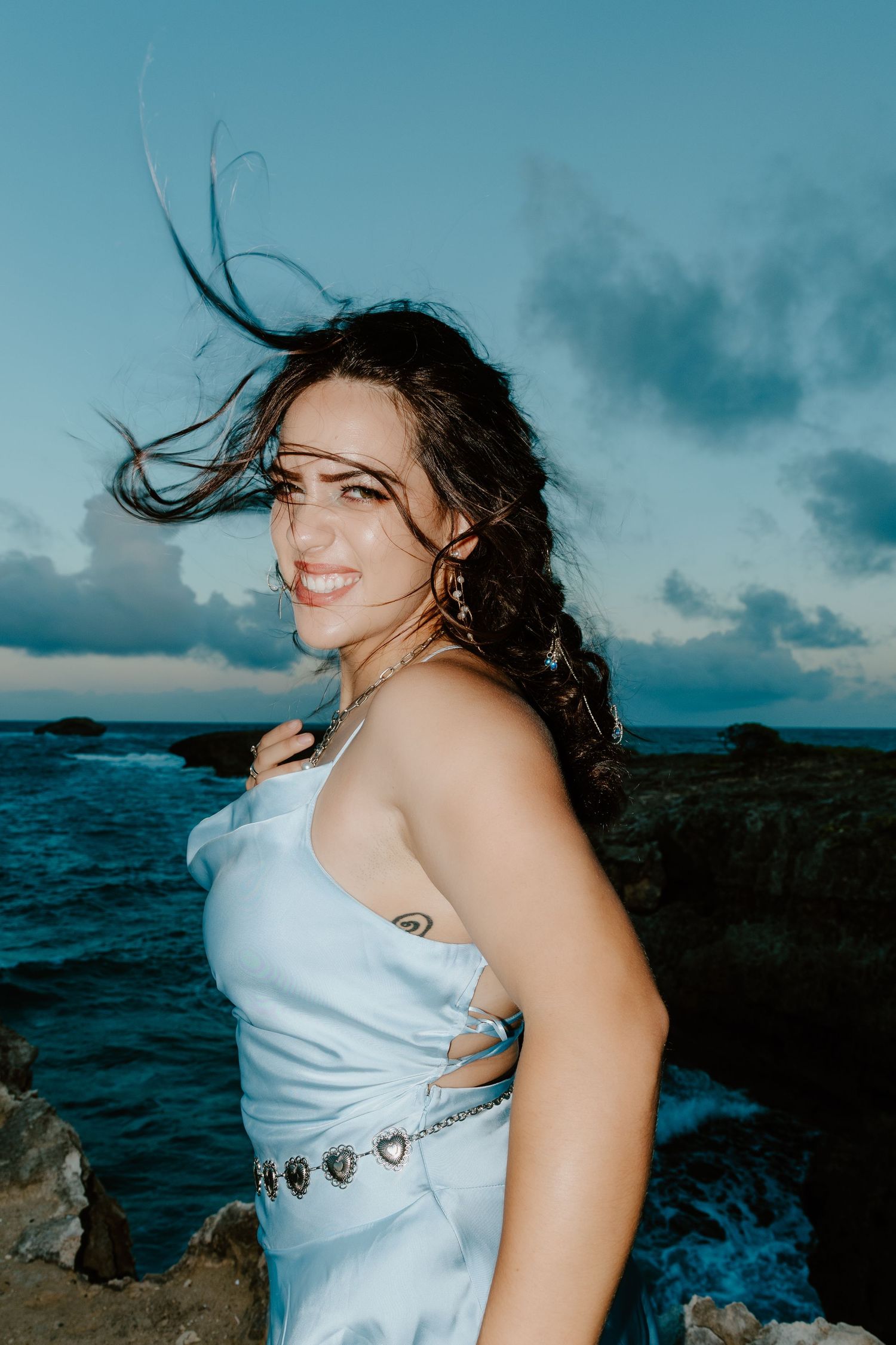 Woman in white dress smiling by ocean at dusk, hair blowing in wind, rocky coastline background.