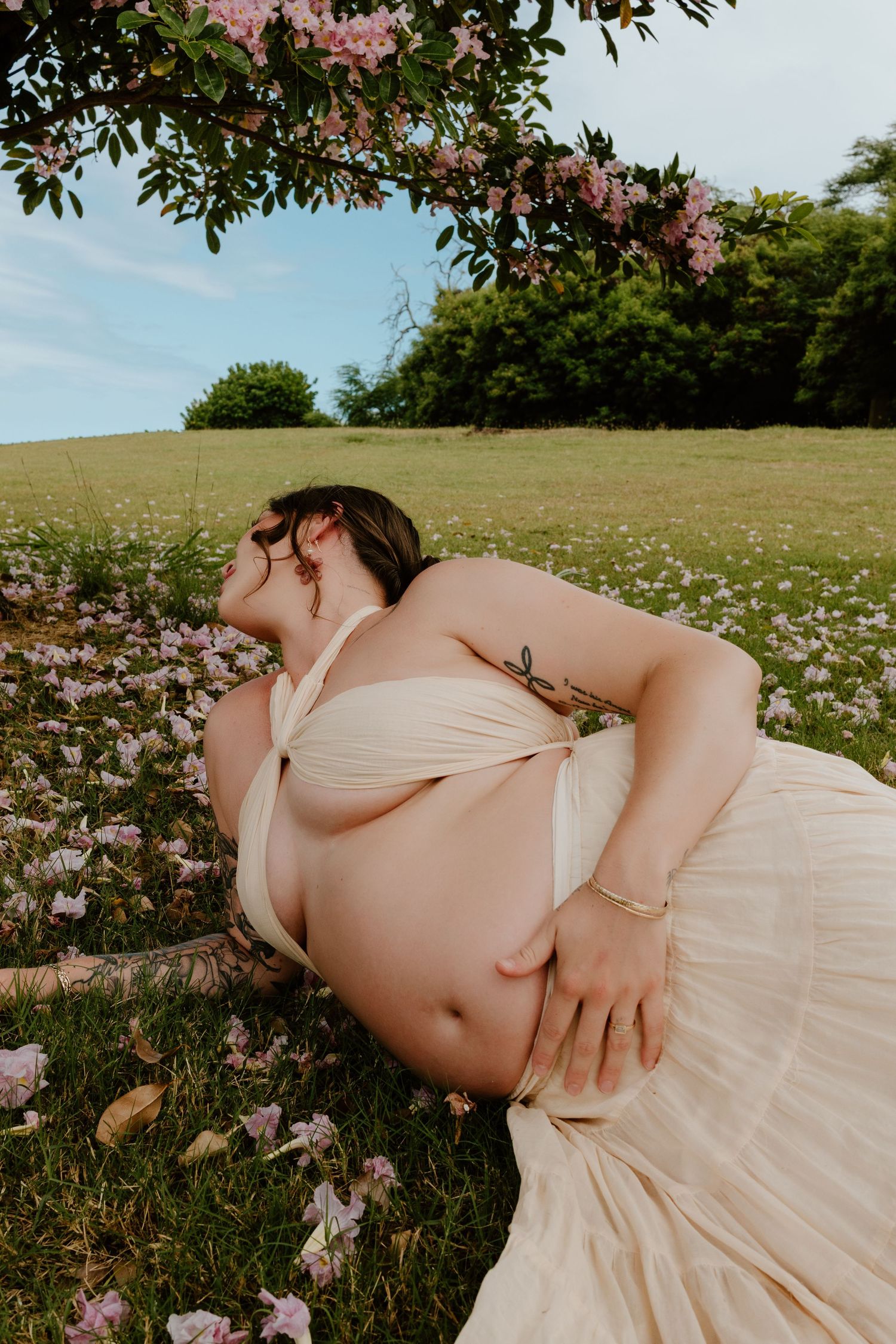 Pregnant woman in cream bikini top and skirt reclines in flower-filled meadow under blooming pink tree.