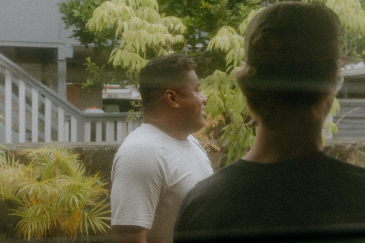 Two men standing outdoors near green tropical plants and a white railing, one wearing a white t-shirt.