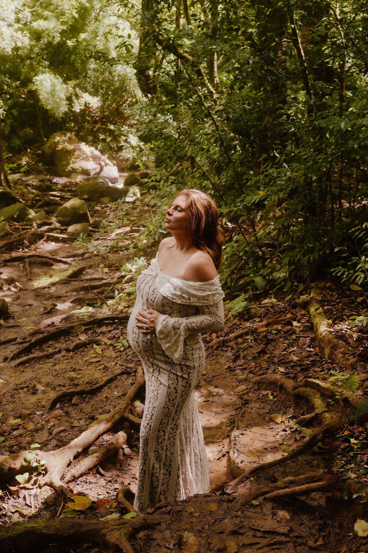 Pregnant woman in elegant lace off-shoulder gown standing on forest path near waterfall.