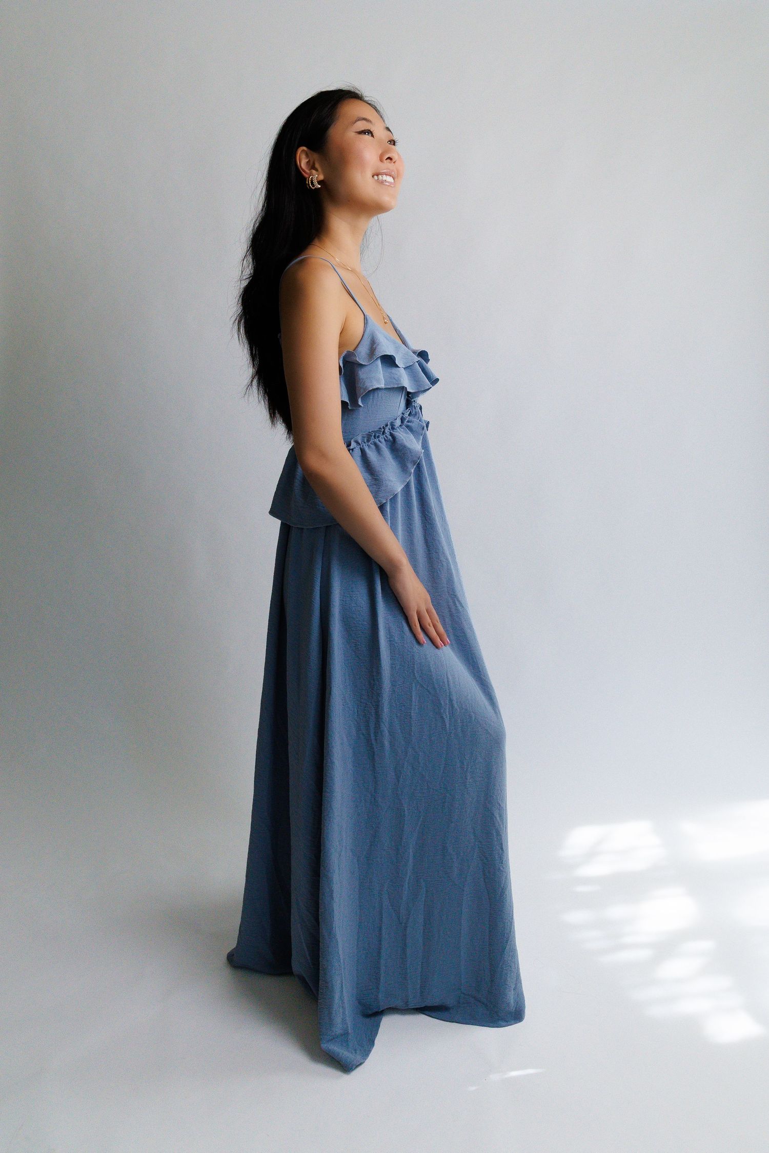 Woman in blue satin maxi dress with ruffle bodice and spaghetti straps, smiling sideways in studio.