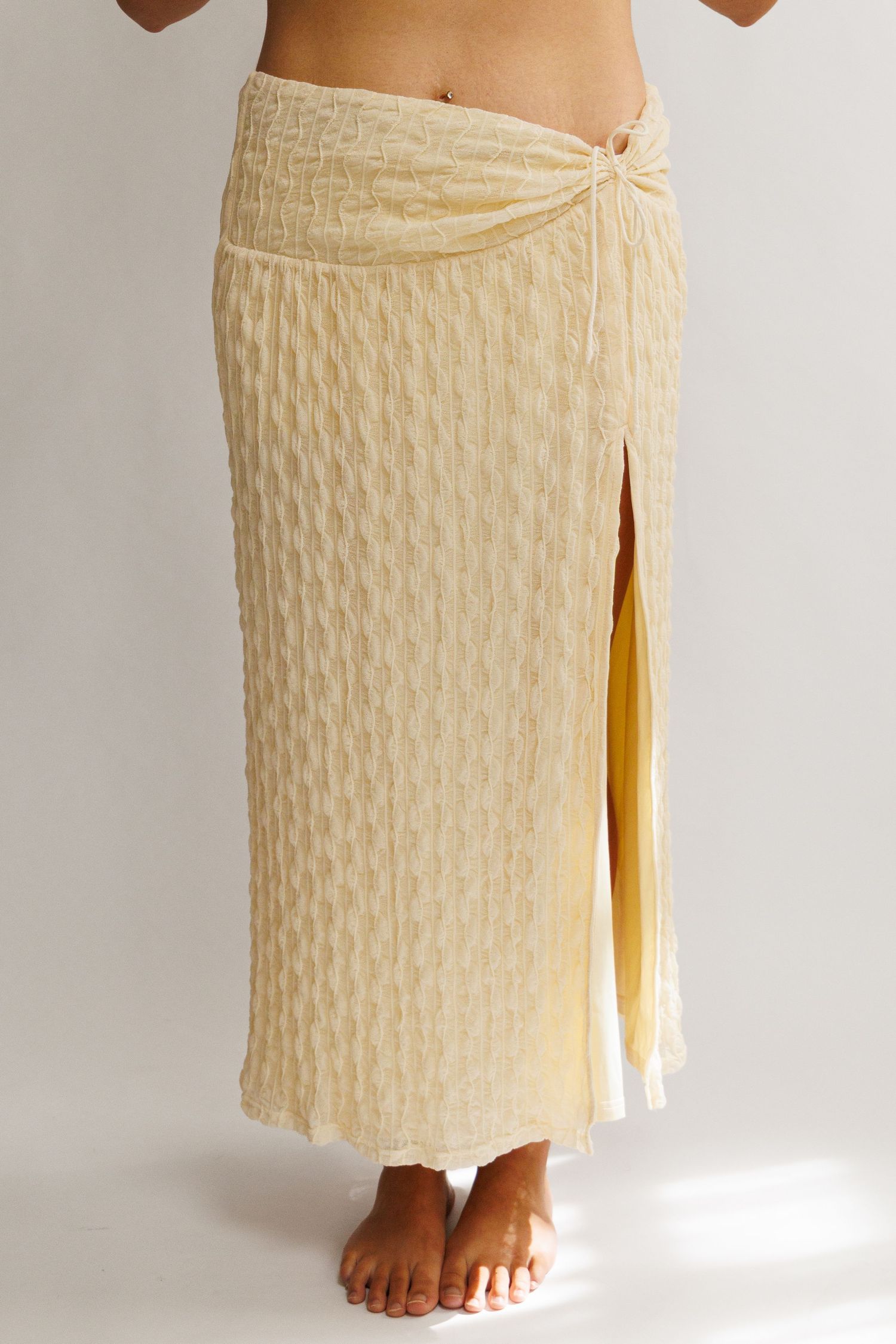 Pale yellow textured midi skirt with front slit and knotted waistband detail, worn by a barefoot model.