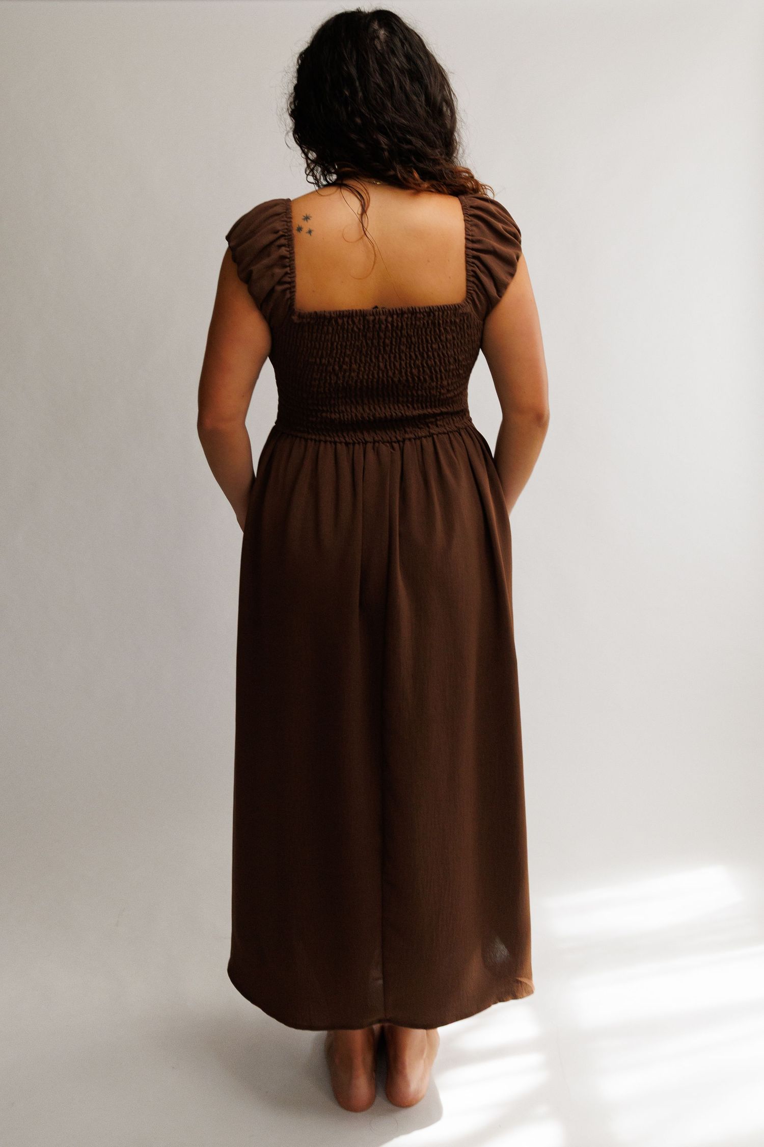 Back view of a woman wearing a brown smocked bodice midi dress with ruffle cap sleeves and pockets.