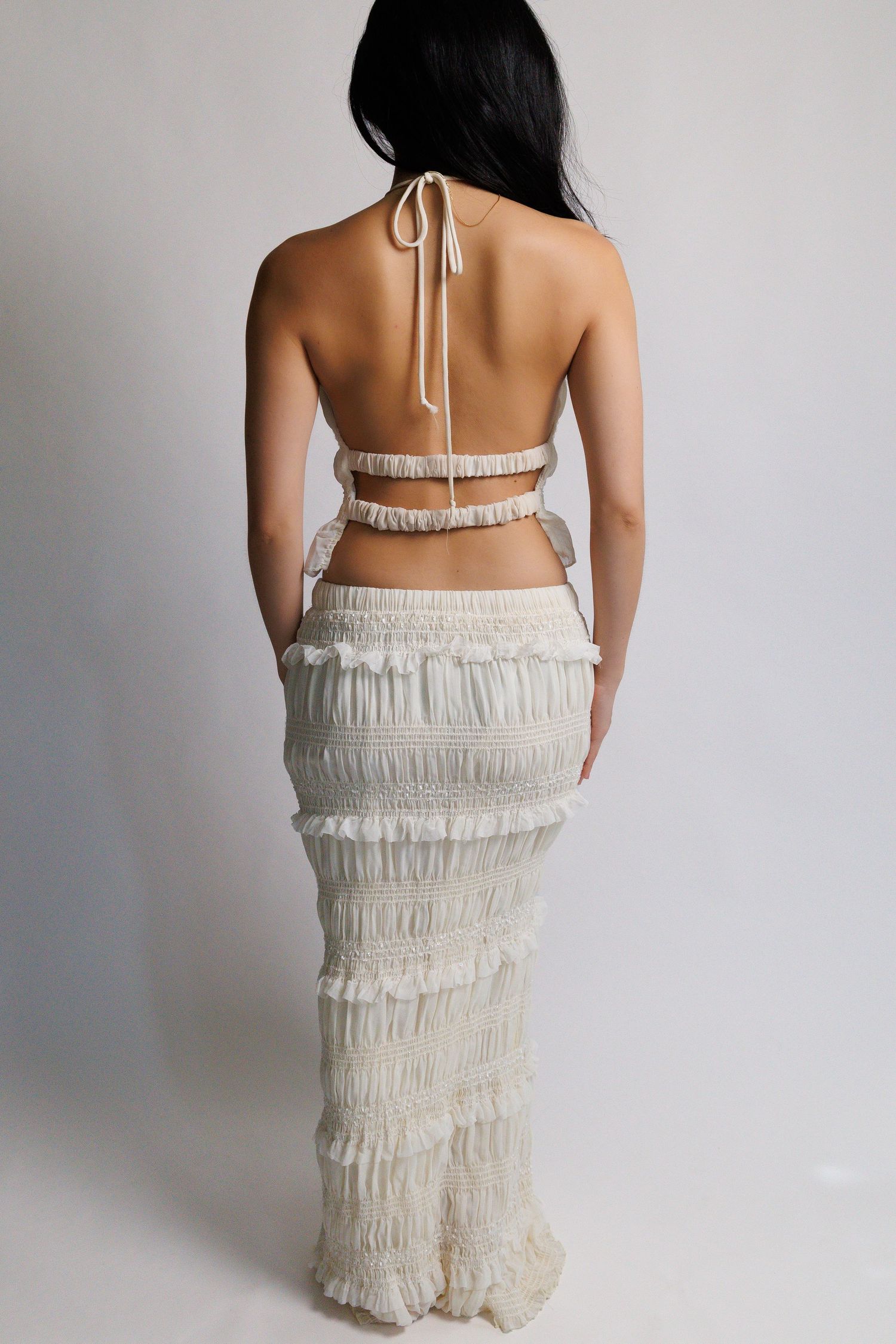 Woman wearing cream ruffled halter crop top and matching maxi skirt set, backless design, photographed from behind.