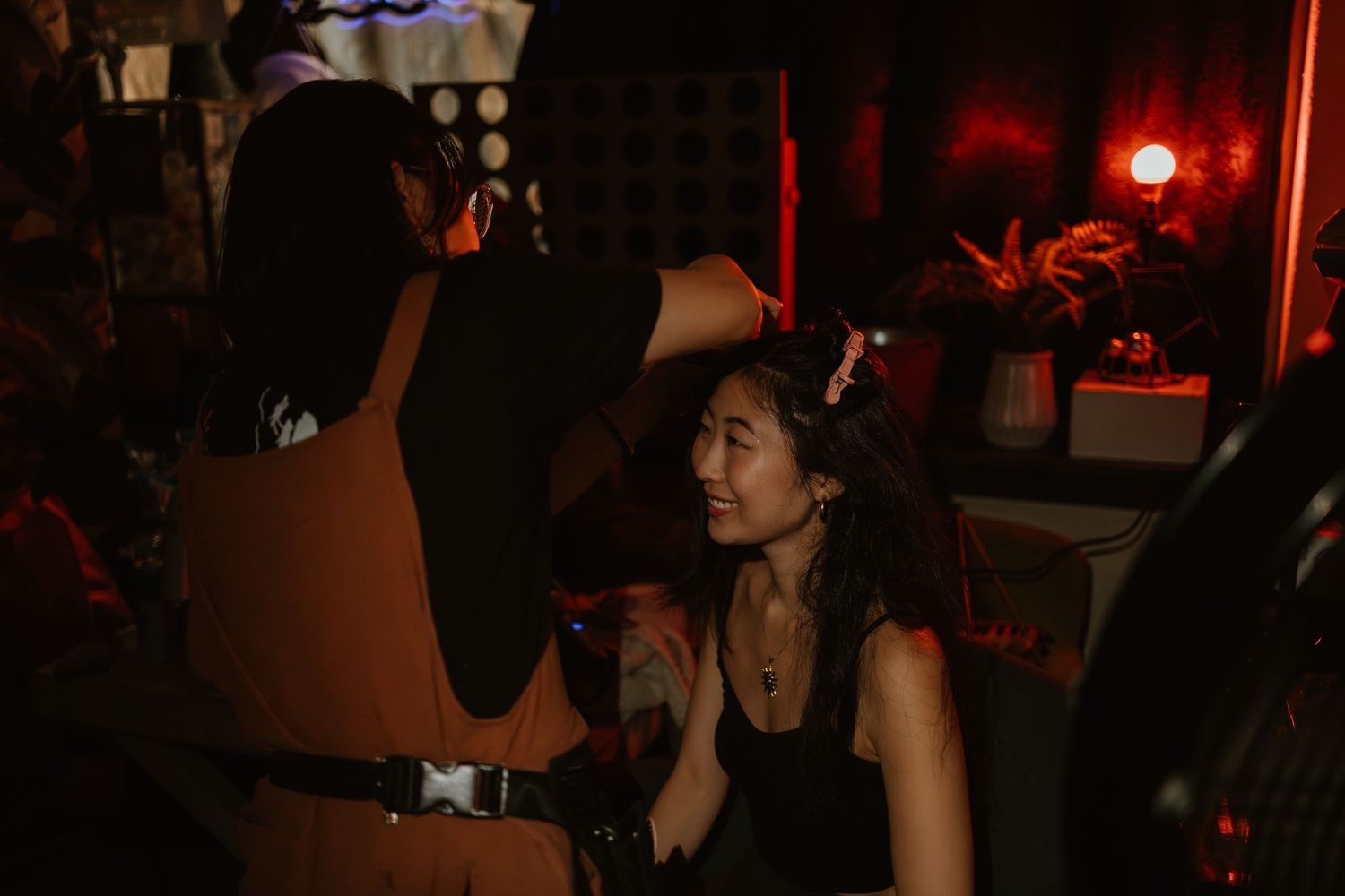 Two women in a dimly lit nightclub, one styling the other's hair under red ambient lighting.