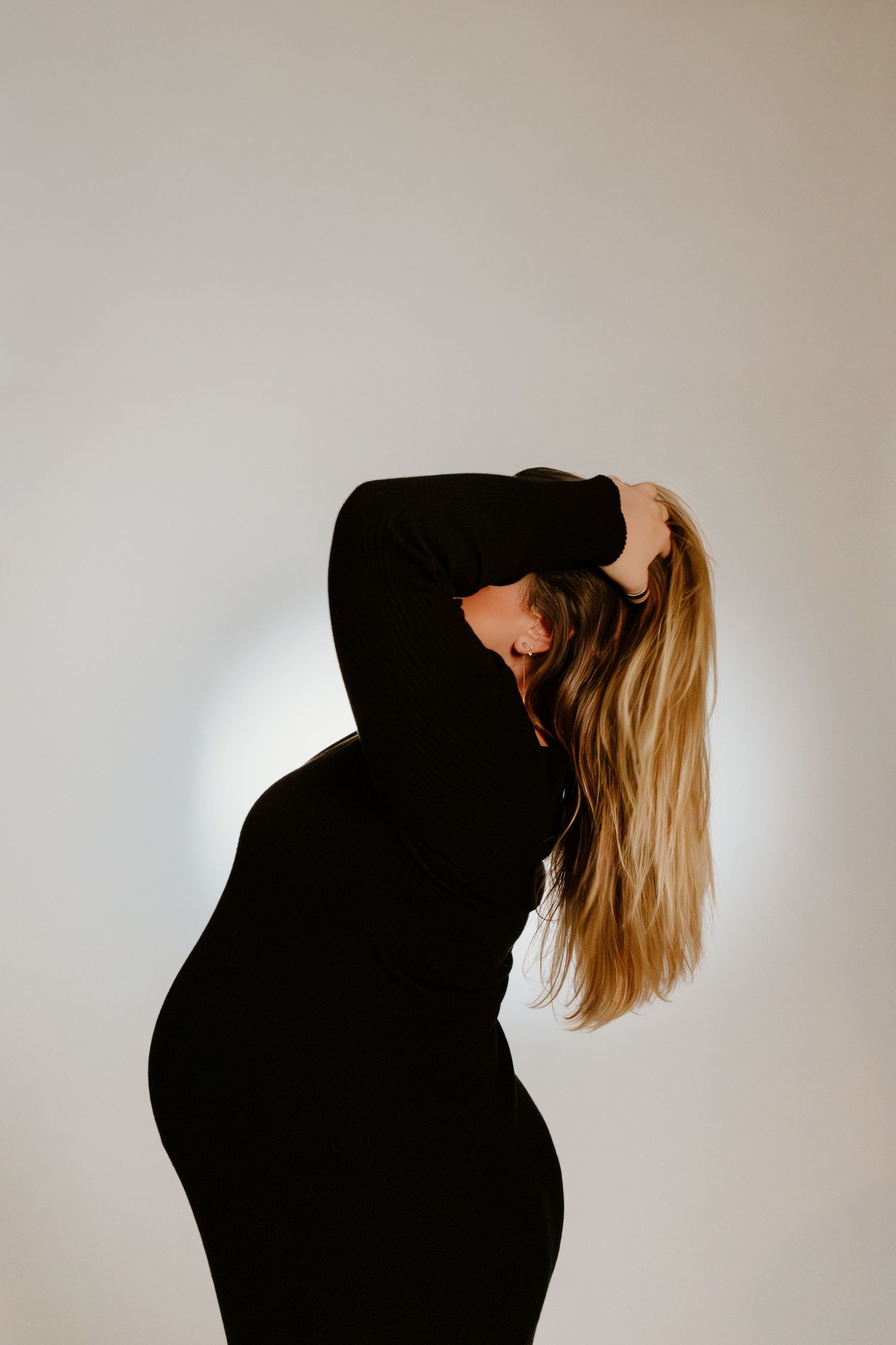 Pregnant woman in black dress pulling hair back, profile silhouette against bright white background.