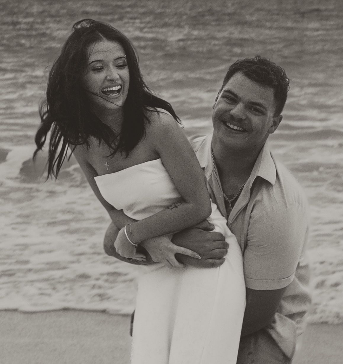 Black and white photo of laughing couple on beach, man lifting woman in white dress near ocean waves.
