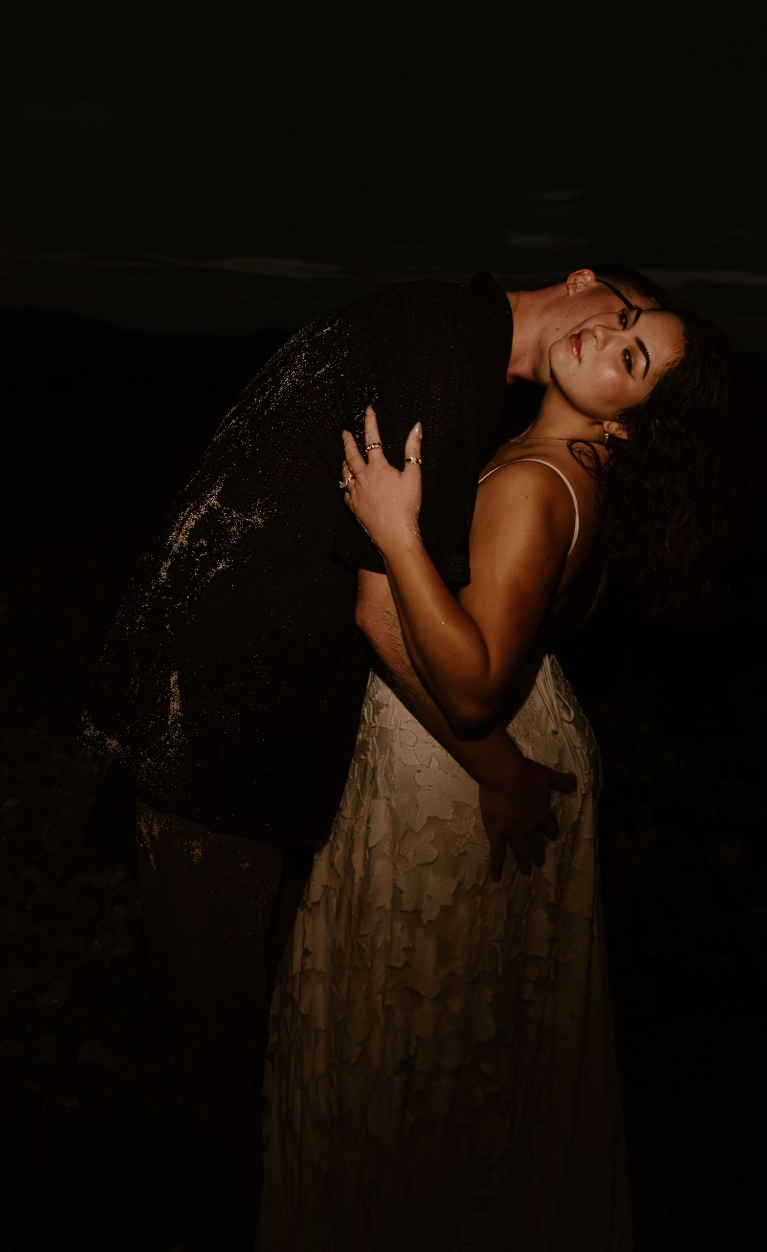 A bride in white lace gown embraces her groom in a dramatic dark nighttime outdoor portrait.